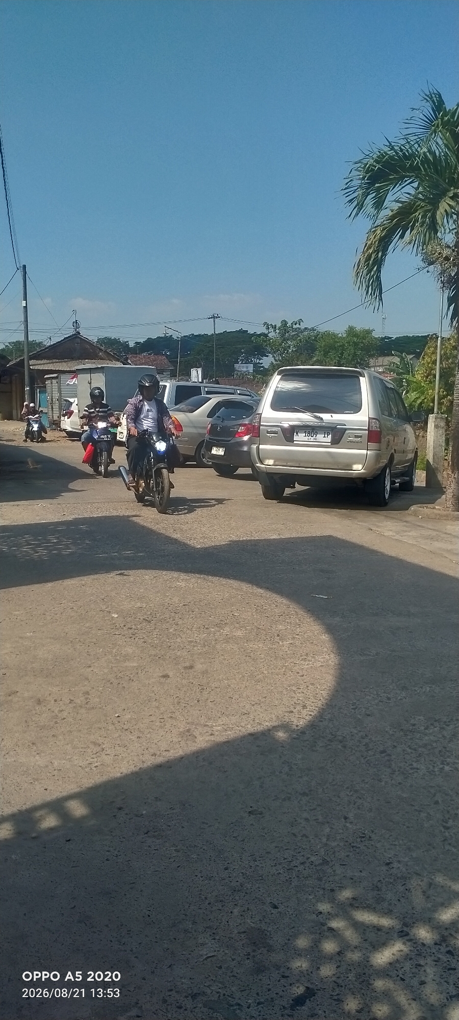 This is a medium-shot, eye-level, vertical photograph of a street scene in Dusun Satu, Indonesia, under a clear blue sky. The foreground is dominated by a rough, sun-drenched asphalt road. The middle ground features several vehicles: a silver SUV with a license plate reading "K 1809 IP" is parked on the right. To its left, a dark-colored car and a silver sedan are also visible. Several motorcycles are in motion. One motorcycle in the center has a rider wearing a helmet and a patterned shirt, with headlights on. Another motorcycle to the left has a rider with a helmet and a striped shirt, also with headlights illuminated. Further left, another motorcycle with a rider in a blue shirt is partially visible. The background shows a row of low-rise buildings with corrugated roofs, some trees, and utility poles. The overall impression is a casual street scene in a rural or semi-urban Indonesian setting, likely during daylight hours.
