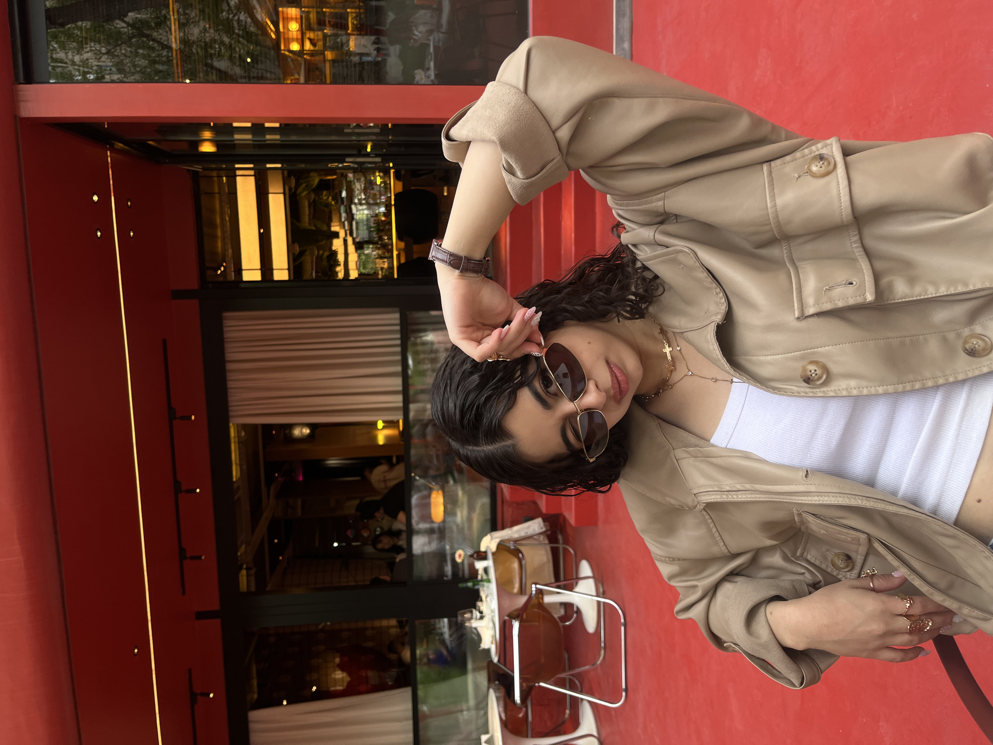 A young woman is posing on a red surface, likely a patio or walkway. She is wearing a beige jacket over a white tank top, sunglasses, and a necklace with a cross. The background reveals a building with a red facade and large glass windows, reflecting interior lights and suggesting an urban environment. The setting seems to be in Yerevan, Armenia, with outdoor seating visible, implying a café or restaurant.