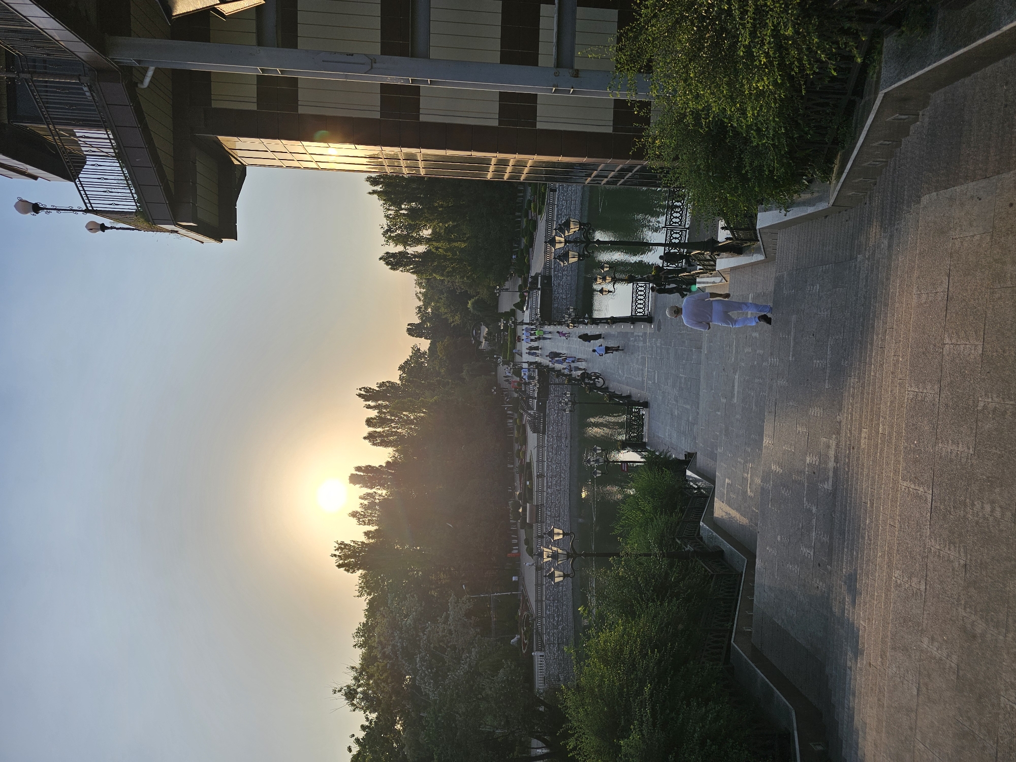 The image shows a view looking down a wide, stone staircase towards a park area. The park features a body of water, a bridge with people walking across it, and trees lining the background. The sun is setting, casting a warm glow over the scene. A modern building with a balcony and glass facade is visible on the right side of the frame. The location is likely in Cherkessk, Russia, possibly near a prominent park or waterfront area, given the architectural style and landscaping.
