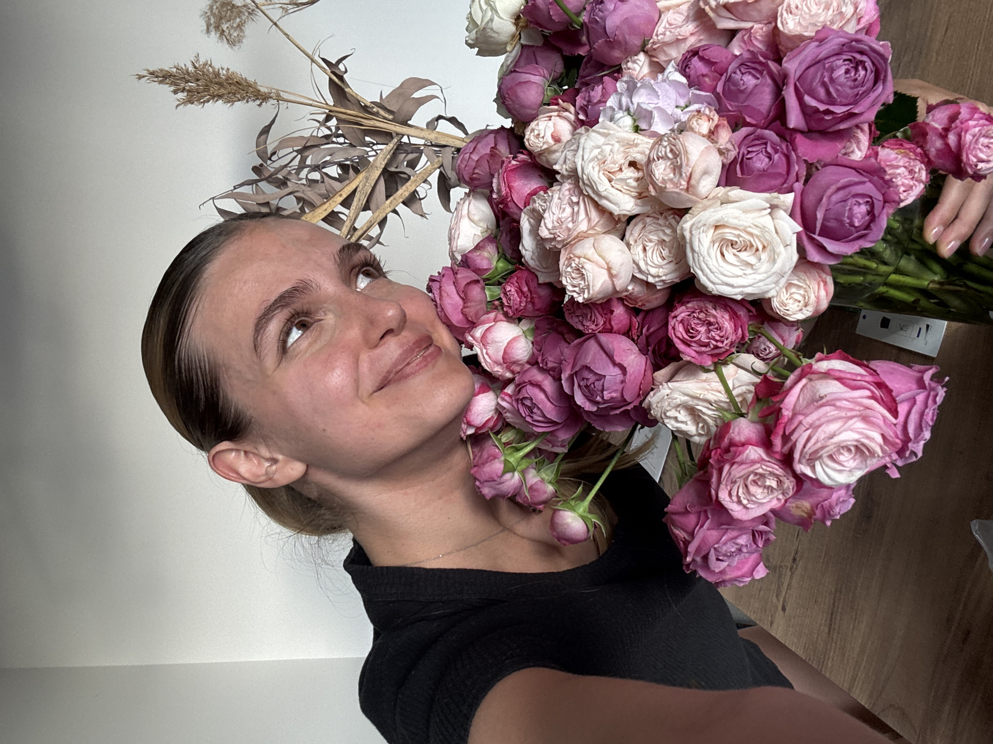 A close-up, slightly rotated, eye-level shot captures a young woman admiring a large bouquet of roses. The woman, positioned on the left side of the frame, has light skin and light brown hair pulled back, revealing her profile. She is wearing a dark, possibly black, ribbed top and a delicate silver chain necklace. Her expression is one of gentle pleasure or admiration, with a subtle smile and eyes gazing upwards towards the flowers.
The substantial bouquet dominates the right and central portions of the image. It consists of numerous roses in various stages of bloom, showcasing a rich palette of pinks, purples, and creamy whites. Some dried floral elements, including feathery reeds and elongated brown leaves, are visible behind and above the woman's head, suggesting a decorative backdrop or additional elements within the arrangement.
The setting appears to be indoors, against a plain, light-colored wall, which serves as a neutral background. A wooden surface, possibly a table or floor, is visible in the bottom right corner. A hand, likely the woman's, is seen at the base of the bouquet, grasping the stems within a clear vase or wrapping. A small white label with the text "VO" is visible near the base of the stems. The lighting is soft and even, highlighting the textures of the flowers and the woman's skin.