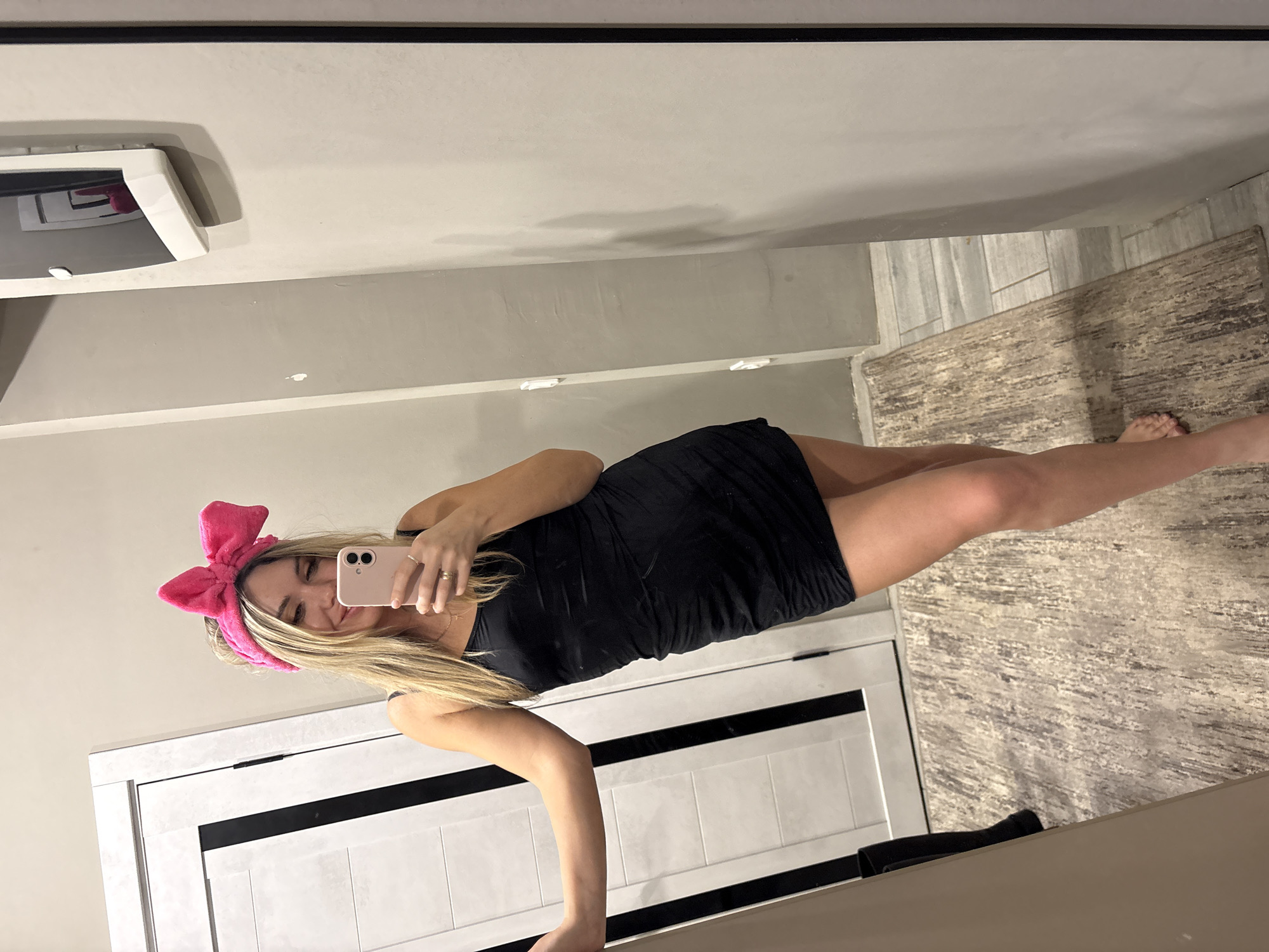 The image displays an adult female posing for a mirror selfie indoors, located in the city of Votkinsk, Russia. The individual has long blonde hair and is wearing a black spaghetti-strap mini-dress, complemented by a large, bright pink, plush bow headband. She holds a light pink smartphone in her right hand, directing it towards the mirror to capture the image; a ring is present on her right ring finger, and a thin necklace with a small pendant is visible around her neck. Her left arm is extended, resting against a white interior door that features black vertical accent strips. She is barefoot and stands on a mottled grey and brown area rug, which partially covers a section of light-colored floor tiles. The walls are painted a neutral light color, exhibiting minor smudges. On the right side of the mirror's reflection, a white, wall-mounted object with a small reflective surface is visible, showing a distorted pink reflection, likely of the headband. A dark, cylindrical object is partially visible on the floor in the lower left quadrant of the frame.