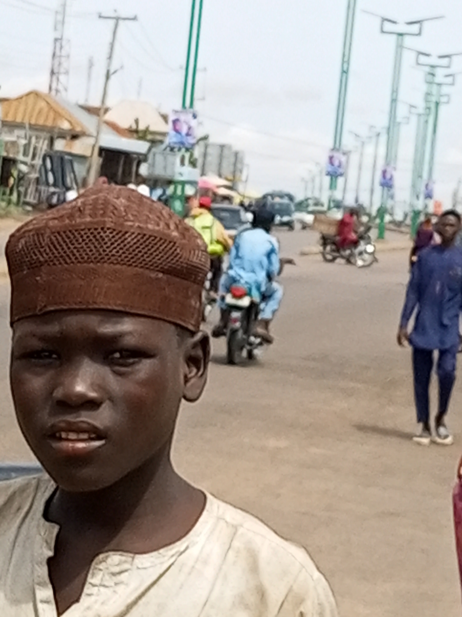 In Garko, Nigeria, on an overcast day, a close-up shot focuses on the face of a young boy wearing a brown, patterned cap. His skin is dark, and his mouth is slightly open, revealing his teeth. He is dressed in a light-colored, collarless garment. The background is blurred, showing a street scene with several people and vehicles. A person in a blue outfit walks towards the right, while another person in a light blue garment rides a motorcycle away from the camera. Several streetlights with solar panels are visible, as are buildings and other objects that are indistinct due to the blur. The overall scene suggests a busy outdoor environment.