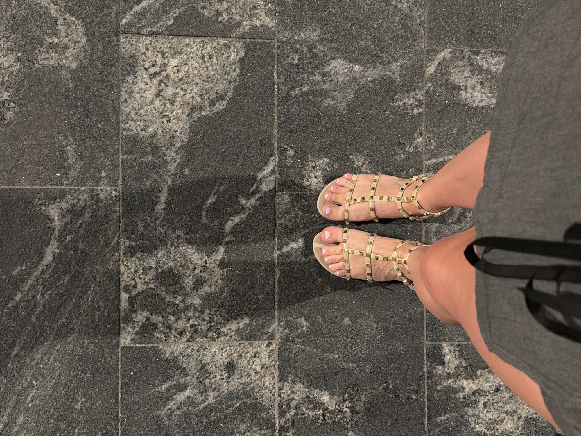A top-down shot shows a person's legs and feet, adorned with gold-studded sandals, standing on a granite-tiled pathway. The tiles are dark gray with lighter gray and white veining. The person is wearing a gray dress, and the sunlight casts shadows on the ground. The scene is set on a bright, sunny day.