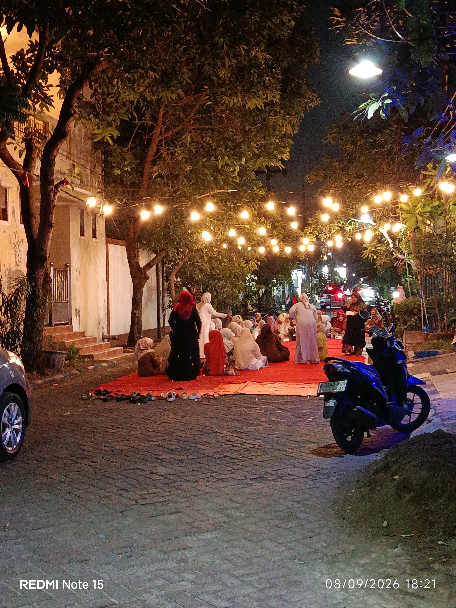 This photograph depicts an outdoor nighttime gathering in Keboansikep, Indonesia, illuminated by strings of warm, festive lights strung between trees. The scene centers on a group of individuals, predominantly women dressed in modest attire, including headscarves, seated on a large red mat spread across a cobblestone street. Some are engaged in conversation, while others appear to be listening or observing. A motorcycle is parked on the right side of the frame, its blue color reflecting the ambient light. In the background, a building with light-colored walls and windows is visible, along with more trees and the suggestion of a street with moving vehicles further down. The overall atmosphere is social and communal, taking place on a paved street that has been temporarily transformed for the occasion.