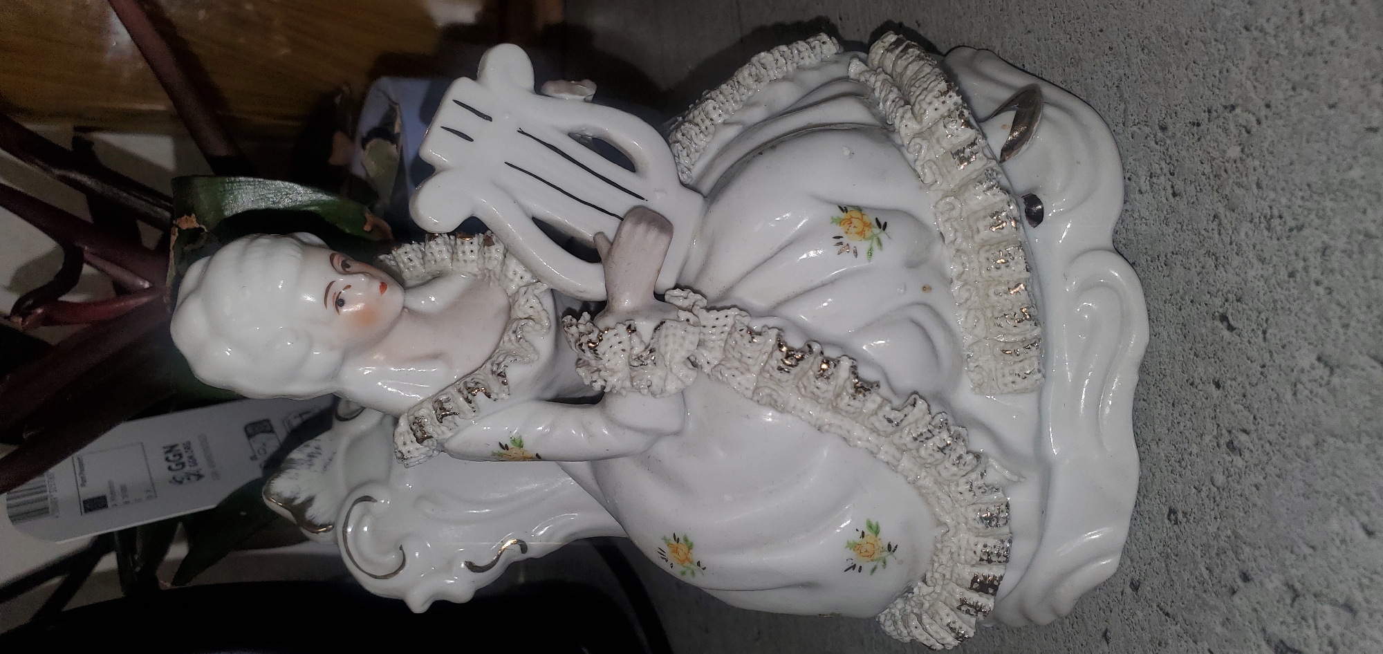 The image shows a close-up of a porcelain figurine of a woman in historical dress, holding a lyre. The figurine is white with delicate floral patterns and gold accents. It is placed on a gray, textured surface, possibly a countertop or table. In the background, there is a blurred plant with dark leaves and a price tag with German text, including "GGN.ORG". The figurine's details, such as her hair, face, and the ruffles on her dress, are clearly visible. The lighting is somewhat dim, casting soft shadows. This image was taken in Sasbach, Germany.