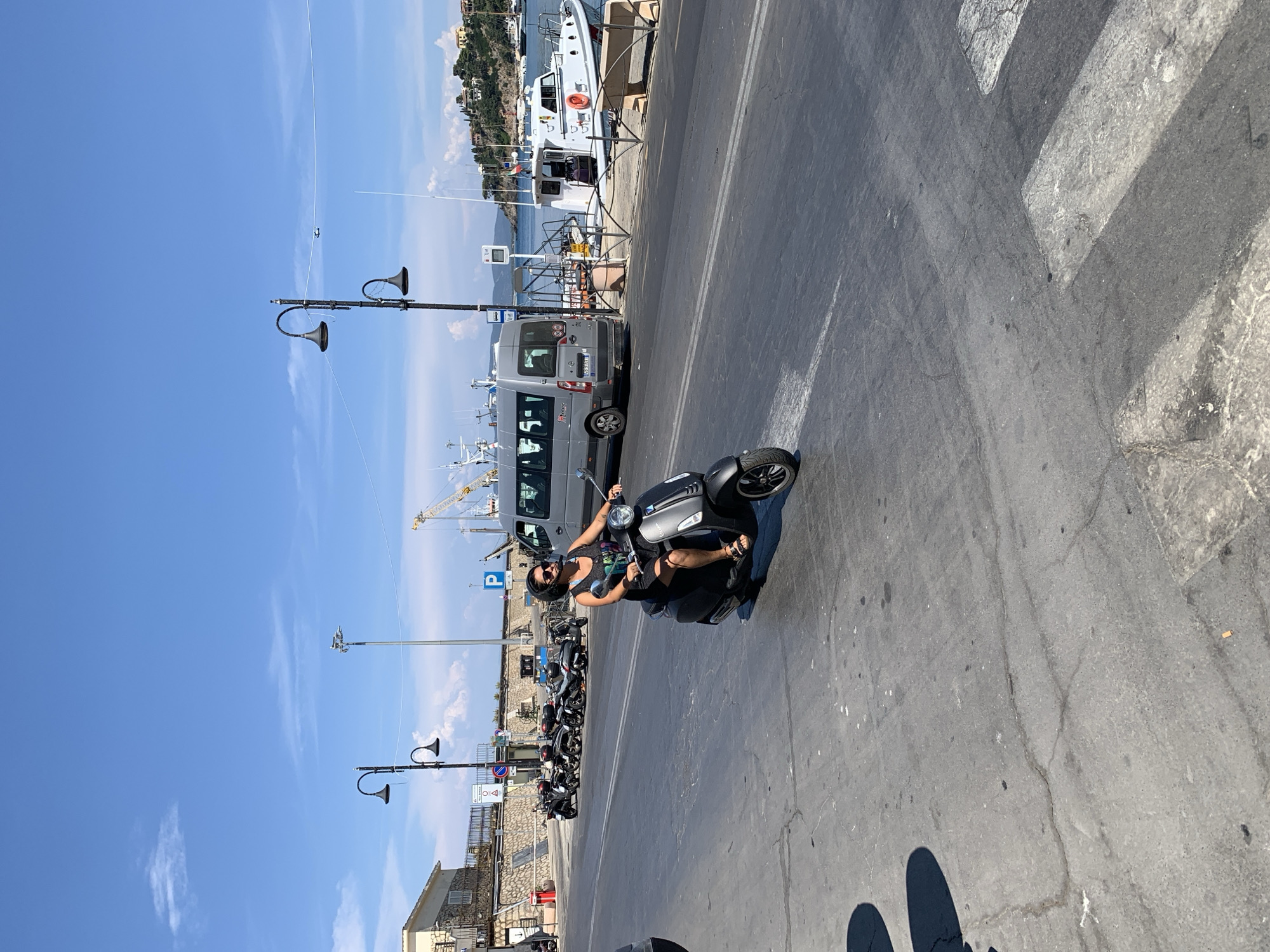 A person is riding a dark-colored scooter on a paved road. They are wearing a black tank top, dark shorts, sunglasses, and sandals. The scooter is a modern design with a headlight and handlebars. The scene is outdoors in a harbor area, with boats docked in the background and a crane visible further down. A grey van is parked behind the scooter, and a row of other parked scooters is visible to the left. The road has a pedestrian crossing painted on it. The sky is bright blue with some light clouds, suggesting a sunny day. The overall mood appears cheerful and relaxed, with the rider smiling and seemingly enjoying the ride. The time of day appears to be daytime, likely late morning or early afternoon. Location cues such as the boats, harbor setting, and the architecture of the buildings in the distance, along with the context provided, point to Porto Santo Stefano, Italy. No legible text is visible on the person or the scooter, but license plates are present on the van.