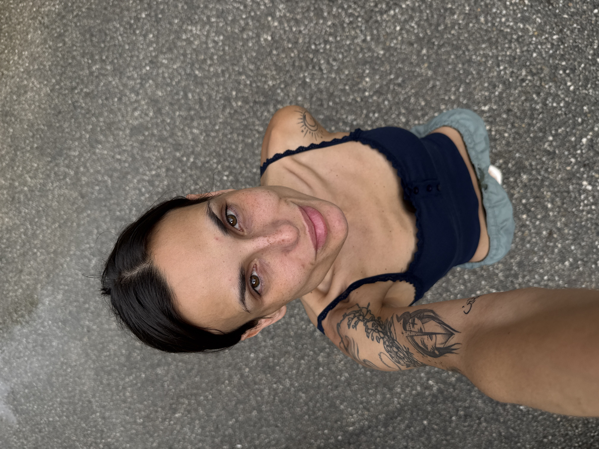 A high-angle, overhead shot captures a woman from the chest up, looking directly upwards at the camera with a gentle smile. She has dark hair styled with a central part, dark brown eyes, and freckles visible across her nose and cheeks. She is wearing a dark blue, spaghetti-strap top with small buttons down the front, and what appear to be light blue or grey bottoms.
Several tattoos are prominent on her arms. On her right shoulder (viewer's left), there is a tattoo depicting a crescent moon with a face and radiating lines, resembling sun rays. On her left arm (viewer's right), a larger, intricate tattoo featuring an eye or a face within a wing-like or abstract design extends down her bicep. Another smaller, symbolic tattoo is visible further down her left forearm.
The background is a blurred, mottled grey surface, consistent with asphalt or concrete, suggesting an outdoor urban or paved setting. The lighting is bright and even, indicating daytime conditions. The woman's expression is relaxed and engaging. The event is a portrait photograph taken in Pleven, Bulgaria. No visible text is present in the image.