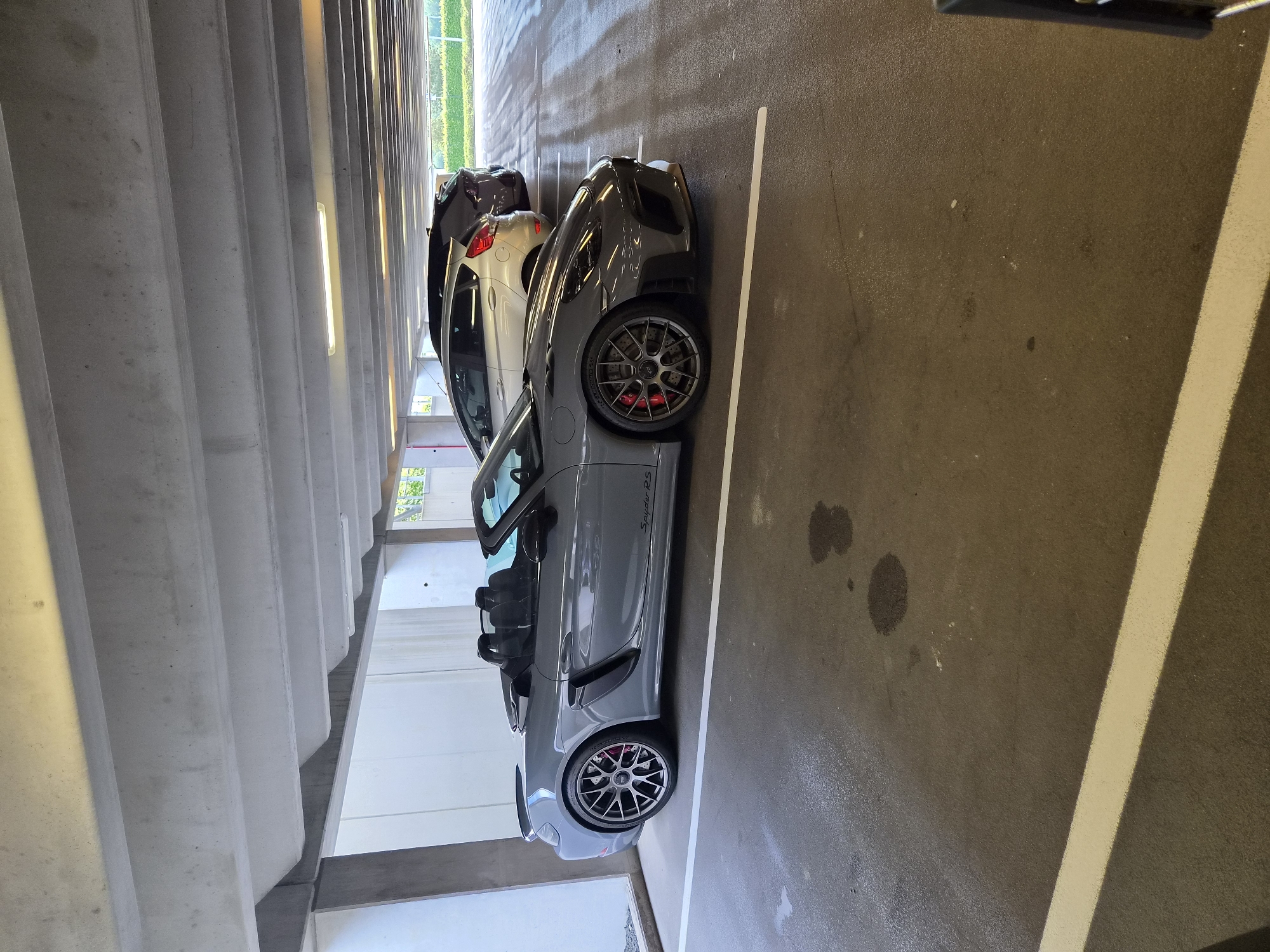 The image depicts a gray Porsche Spyder RS parked in a parking garage. The location is likely Sanem, Luxembourg. The car is parked in a designated spot marked by white lines on the asphalt. There are other cars parked nearby. The environment is a typical parking garage with concrete pillars and a concrete ceiling. There are no people visible in the image.