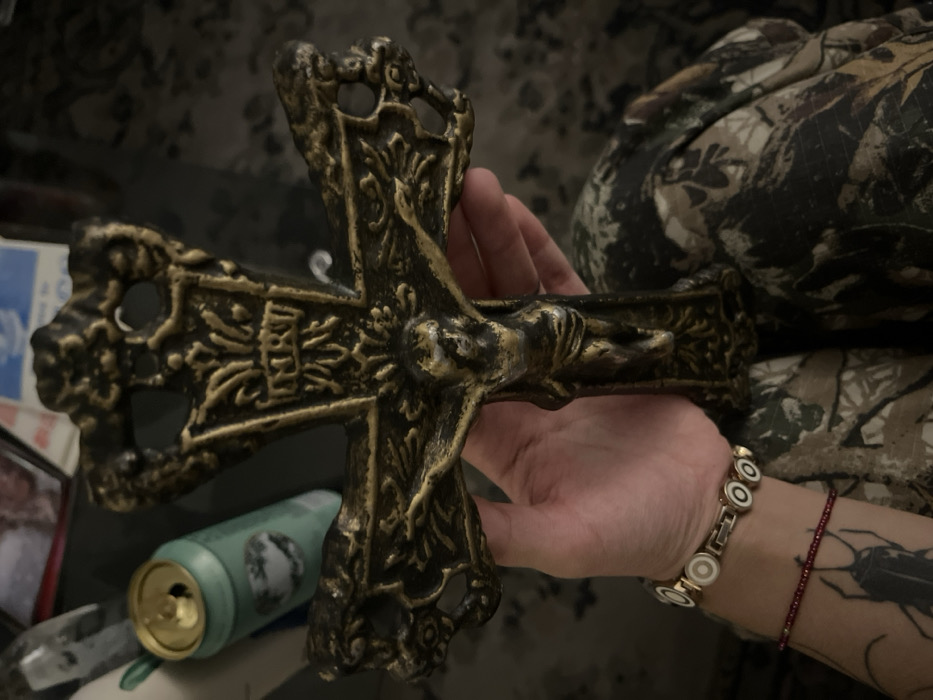 A hand holds an ornate, antique-looking crucifix with a sculpted figure of Jesus. The cross is metallic, possibly brass or bronze, with detailed carvings. A beer can rests in the background along with some miscellaneous items. The person's arm is visible, adorned with a bracelet. A tattoo of a bug is also visible on the forearm. The background consists of a camouflage patterned fabric.