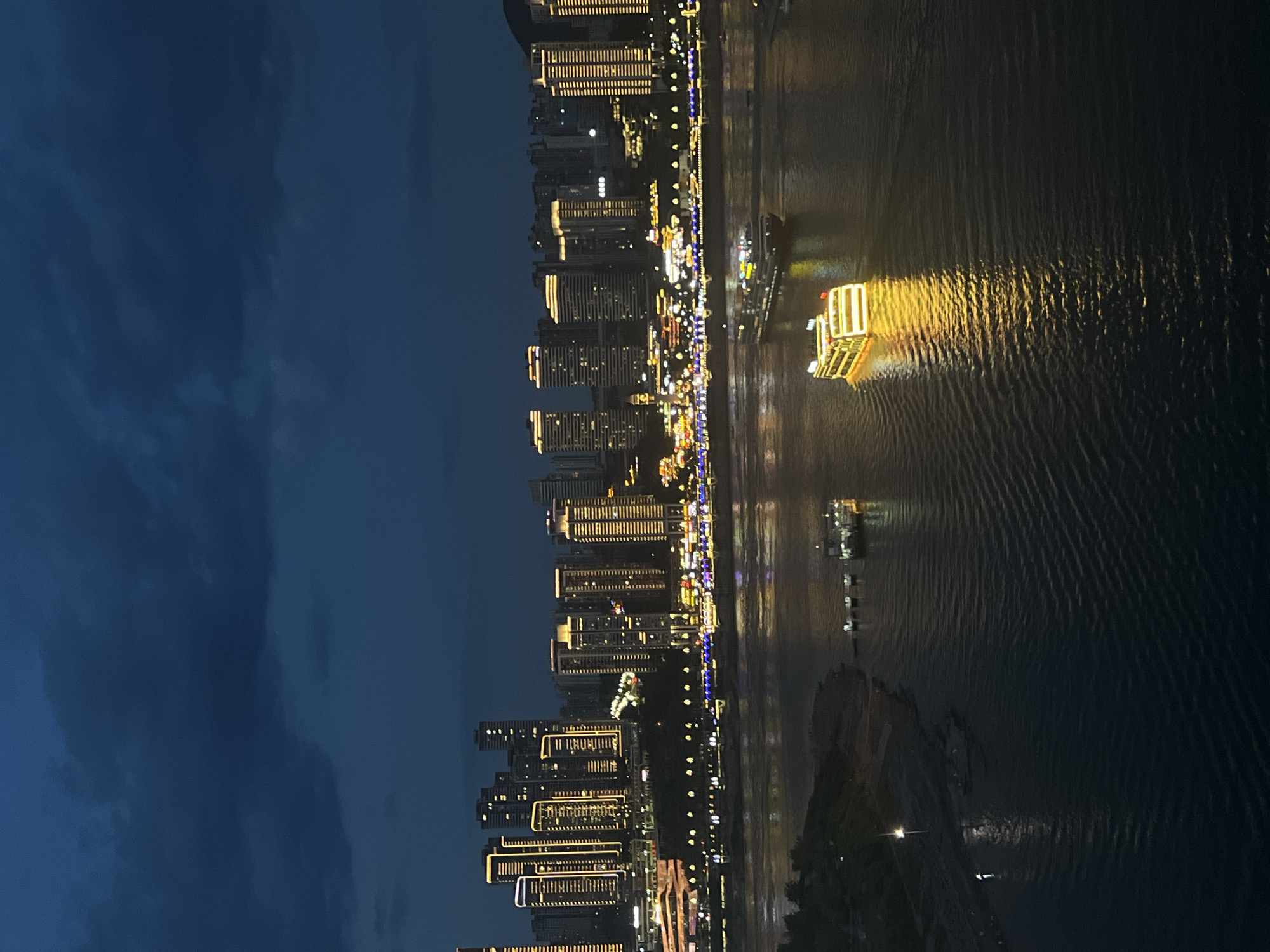 The image captures a nighttime cityscape in Yuzhong District, China, with a wide river in the foreground and illuminated skyscrapers lining the opposite bank. The sky is dark blue with visible clouds. On the river, two boats are present. One boat, closer to the viewer, is brightly lit with yellow lights and appears to be a passenger vessel. Further upstream, another, larger boat with multiple lights is also visible. The river's surface is rippled, reflecting the lights from the city and the boats. The cityscape features numerous tall buildings with yellow and white lights, indicating a well-developed urban area. Along the riverbank, there are additional lights and structures, suggesting a vibrant waterfront. The overall scene depicts a bustling metropolitan area at night, highlighting the contrast between the dark sky and the illuminated urban landscape and river.