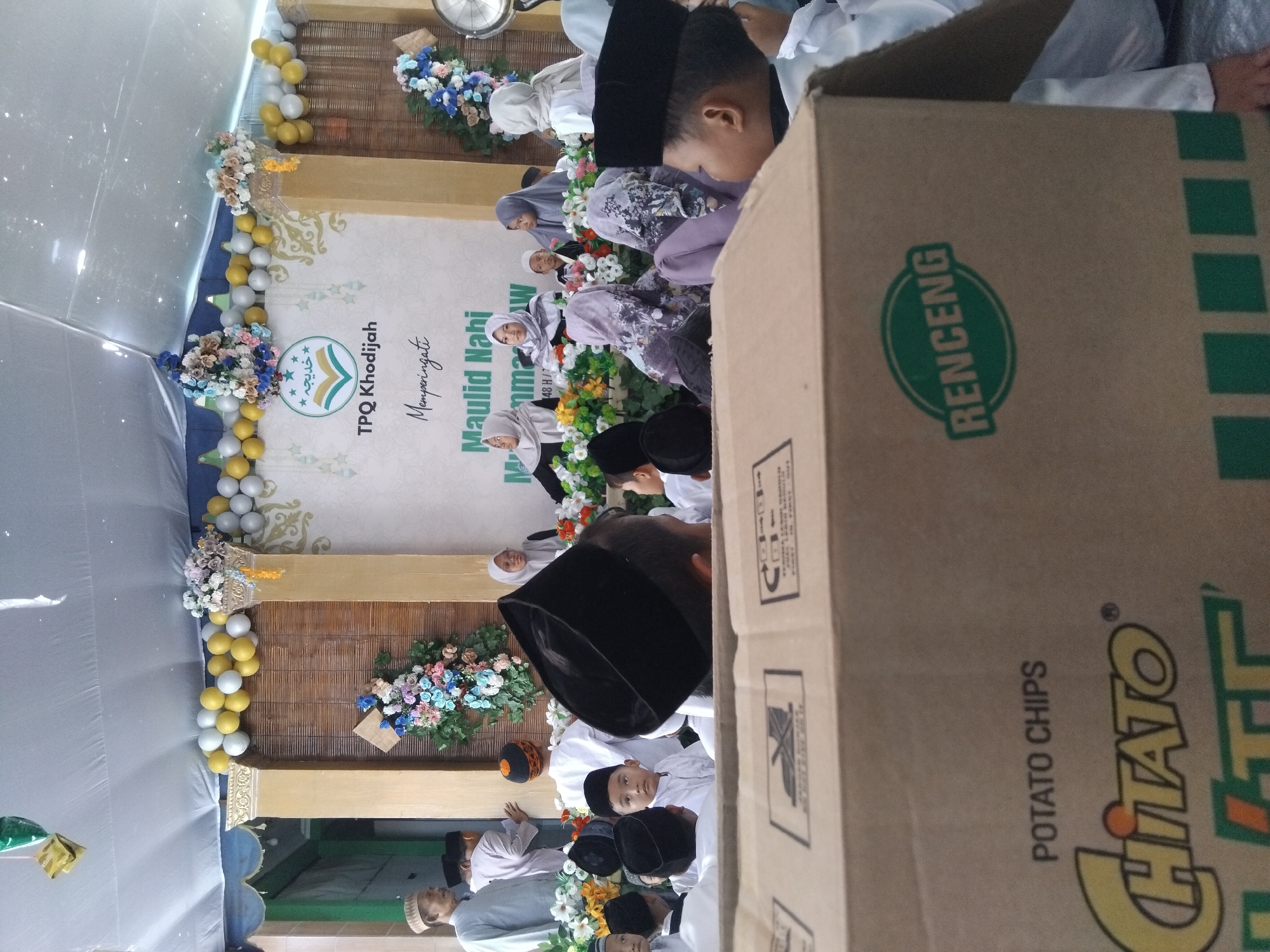 The image captures an indoor event, likely a celebration or ceremony, taking place in a tent structure. The foreground is dominated by a large cardboard box, with the branding "Chitato Potato Chips" and a green emblem reading "RENCENG" visible on its side. In the background, a group of people, primarily children and a few adults, are gathered. Many of the children are wearing white clothing and skullcaps, suggesting a religious or educational context. Some adults are also present, some wearing headscarves. A stage area is set up at the back, decorated with balloons and floral arrangements. A banner with Arabic text and the words "TPQ Khodijah" and "Maulid Nabi Muhammad" is visible on the stage, indicating the event is a commemoration of Prophet Muhammad's birthday at the TPQ Khodijah. The overall atmosphere appears to be festive and communal. The specific location is identified as being in the city of Kepuh Kiriman, Indonesia.