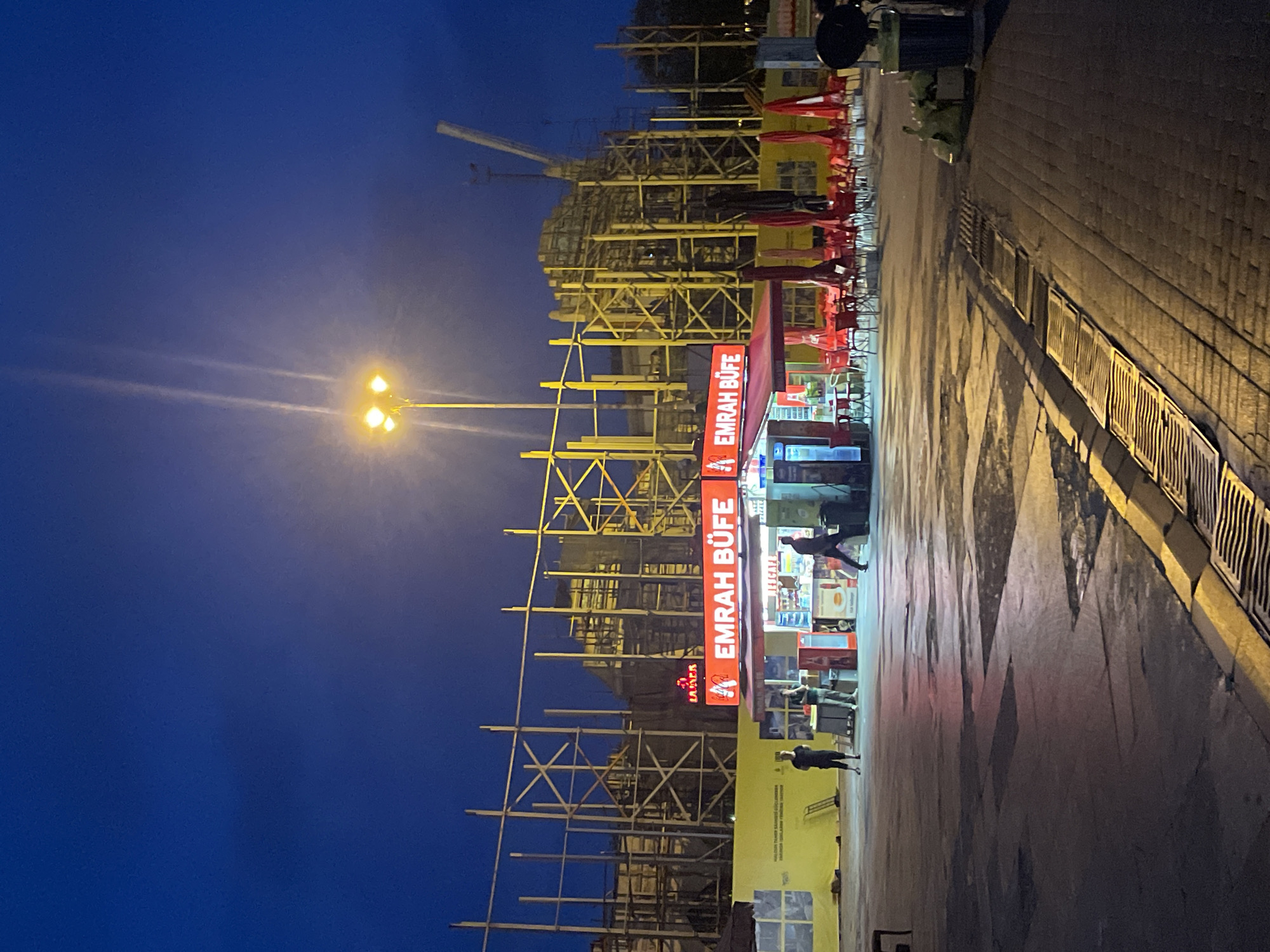 The image depicts an outdoor scene at dusk or early night in Kadıköy, Turkey, with a clear blue sky overhead. In the foreground, a paved area shows reflections of light from the surrounding environment. A sidewalk with cobblestone patterns and a metal grate is visible on the right. In the mid-ground, a brightly lit establishment with a red awning and illuminated signage reading "EMRAH BUFE" is prominent. This establishment appears to be a food vendor or small cafe with red chairs and tables set up outside, some with umbrellas. To the left of the bufe, two individuals stand near a yellow barrier. Behind the bufe and extending across the background is a large-scale construction site characterized by extensive wooden scaffolding and some visible building structures and a crane. The overall atmosphere suggests an urban environment experiencing ongoing development.