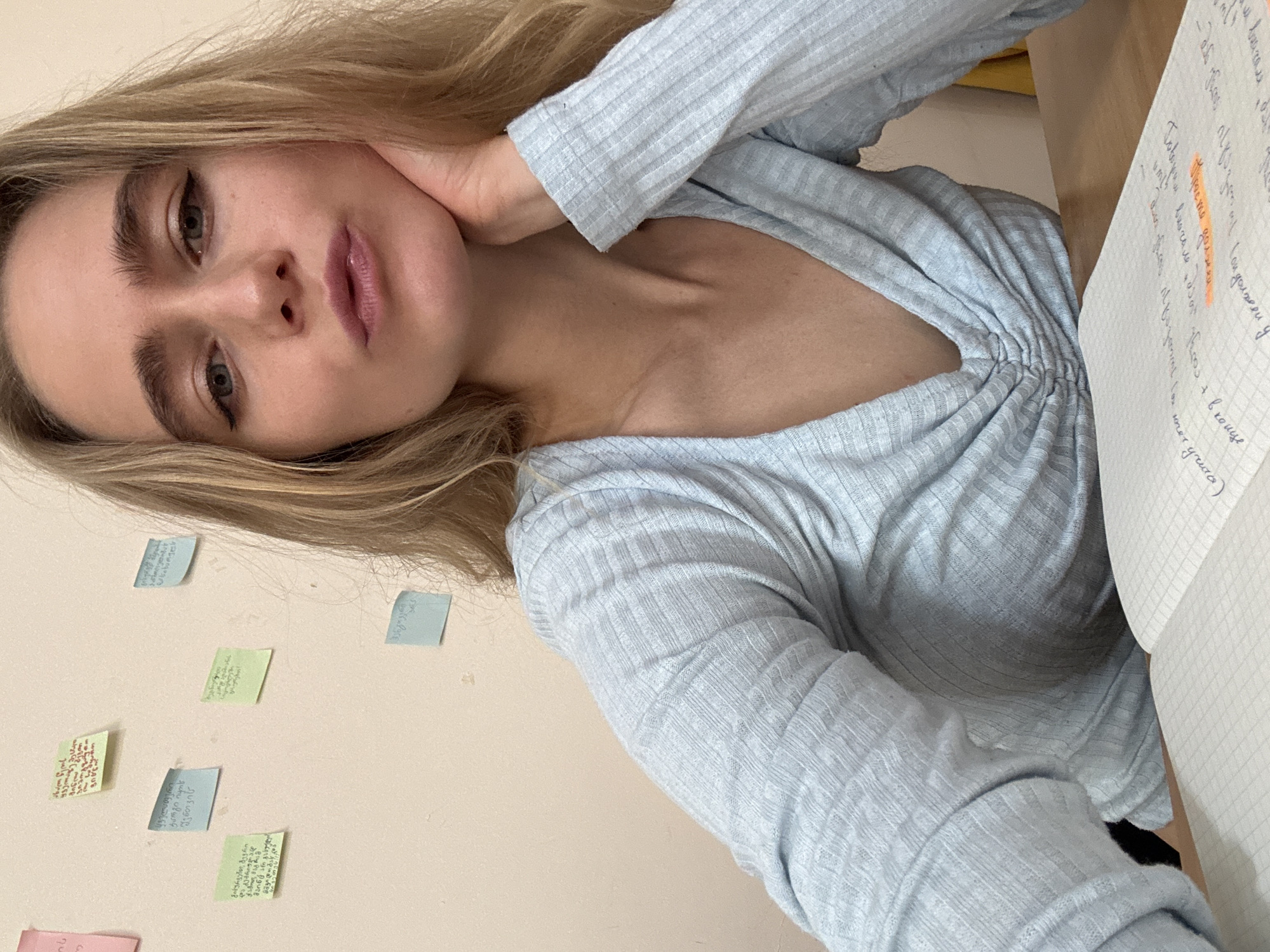 A woman with blonde hair, wearing a light blue ribbed shirt, is looking at the camera with her hand near her neck. There is a notebook with handwriting on it to her right, and various colored sticky notes are adhered to the beige wall behind her.