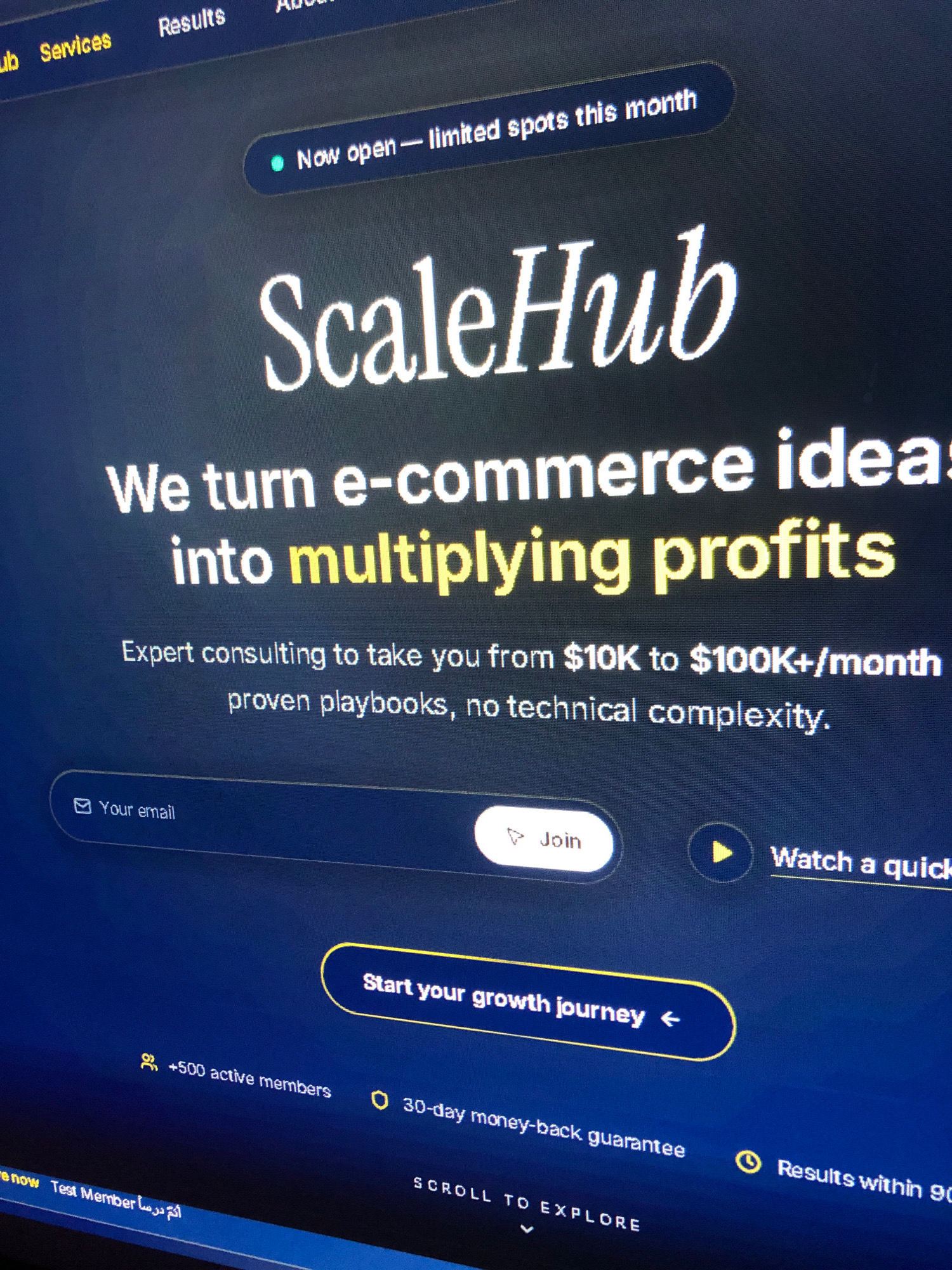 The image shows a computer screen displaying a website for "ScaleHub". The website's tagline is "We turn e-commerce ideas into multiplying profits". Below the tagline, there is a section for users to enter their email and join. There are also buttons for "Join" and "Watch a quick tour". Further down, there is a button that says "Start your growth journey" and text indicating "+500 active members" and "30-day money-back guarantee". At the bottom of the screen, there is text that reads "SCROLL TO EXPLORE". The screen has a dark blue background with white and yellow text. The lighting suggests the room is dimly lit, possibly at night. There is no discernible location within the city of Ali Mendjeli, Algeria, depicted or suggested in this image.