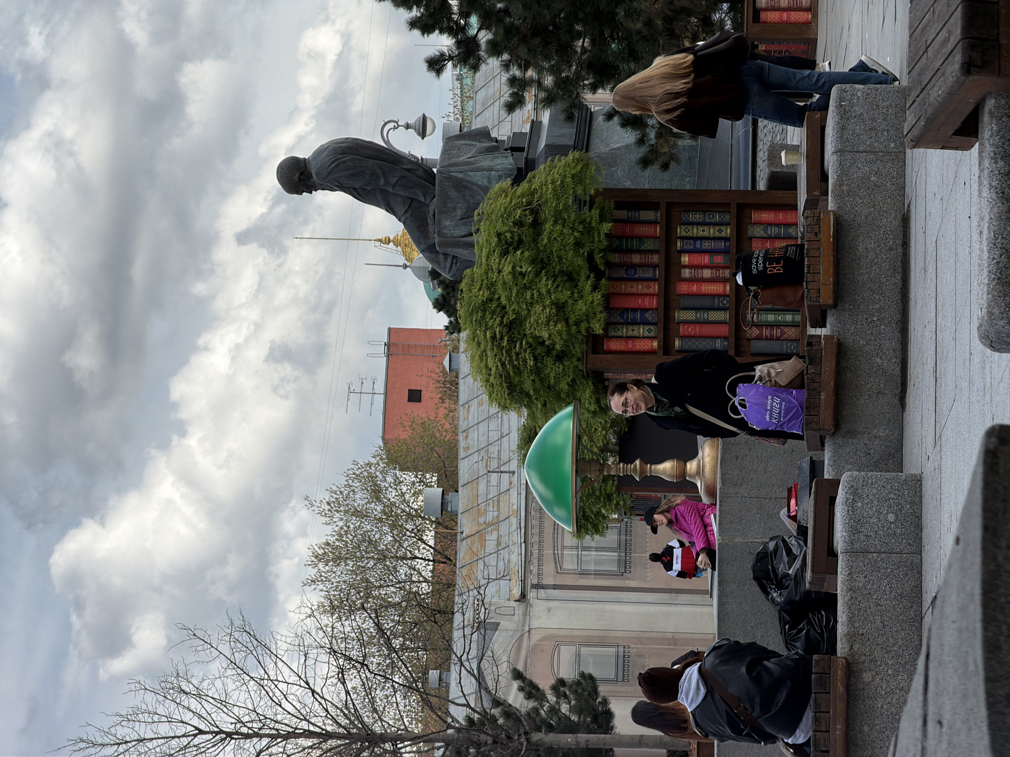 The image captures an outdoor urban scene, likely a public square or pedestrian area, under an overcast sky during daylight hours.
In the left half of the frame stands a large, dark-colored statue of a man seated with his head bowed, positioned on a substantial pedestal. Green bushes surround its base. Behind the statue, a golden dome, possibly part of a church or significant building, is partially visible, along with a reddish-brown building further in the background.
To the right of the statue, a prominent wooden structure designed as a large outdoor bookshelf is filled with numerous books, displaying varied spines in colors like red, blue, and gold. This suggests an open-air book display or a decorative art installation related to literature.
Several people are present in the scene. A woman with long blonde hair, wearing a dark jacket and blue jeans, walks away from the bookshelf area. Closer to the foreground, a woman in a black coat and glasses stands near the book display, holding a purple shopping bag. Nearby, another person in a pink jacket is partially visible. In the lower right corner, several individuals are seen from behind, sitting on stone benches with wooden seats, suggesting a moment of rest or observation.
Visible text includes "КНИГИ" (Kni-gi), meaning "BOOKS" in Russian, printed on the purple shopping bag. On a dark bag placed on a bench near the book display, the English phrases "somebody needs" and "BE HAPPY" are legible. Bare tree branches suggest either late autumn or early spring. The overall atmosphere is that of a calm day in a bustling public space.