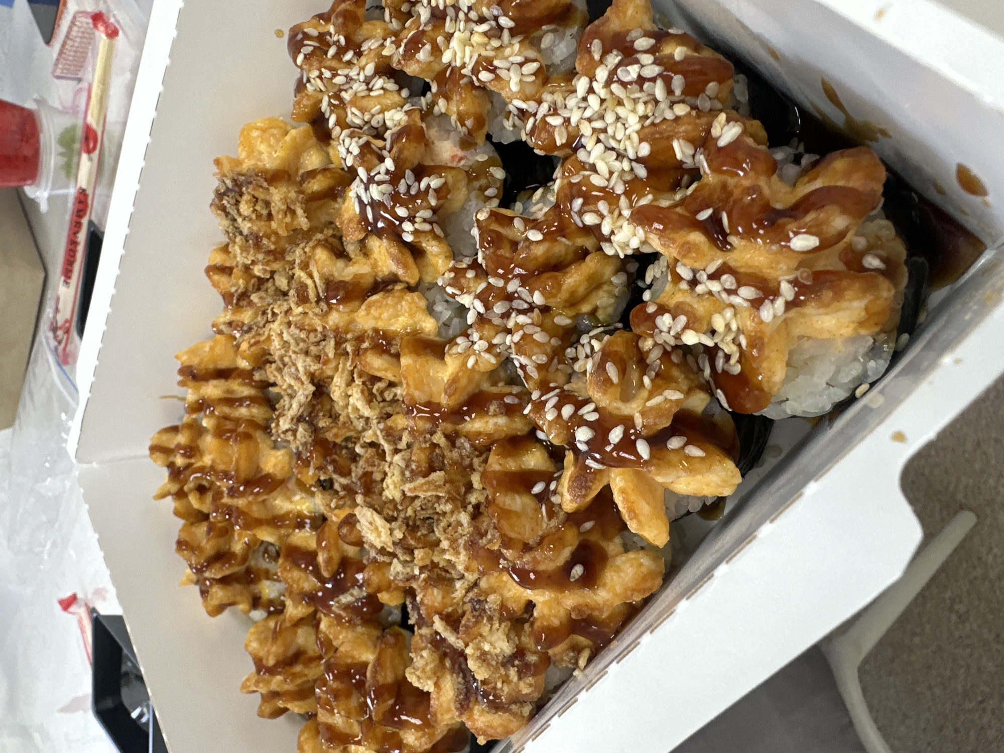 A box of sushi, likely takeout, is displayed in a white container. The sushi appears to be of various types, some with fried toppings and others with sesame seeds, all drizzled with a brown sauce. The image quality does not allow for identifying the specific location in Votkinsk, Russia. No people are visible.