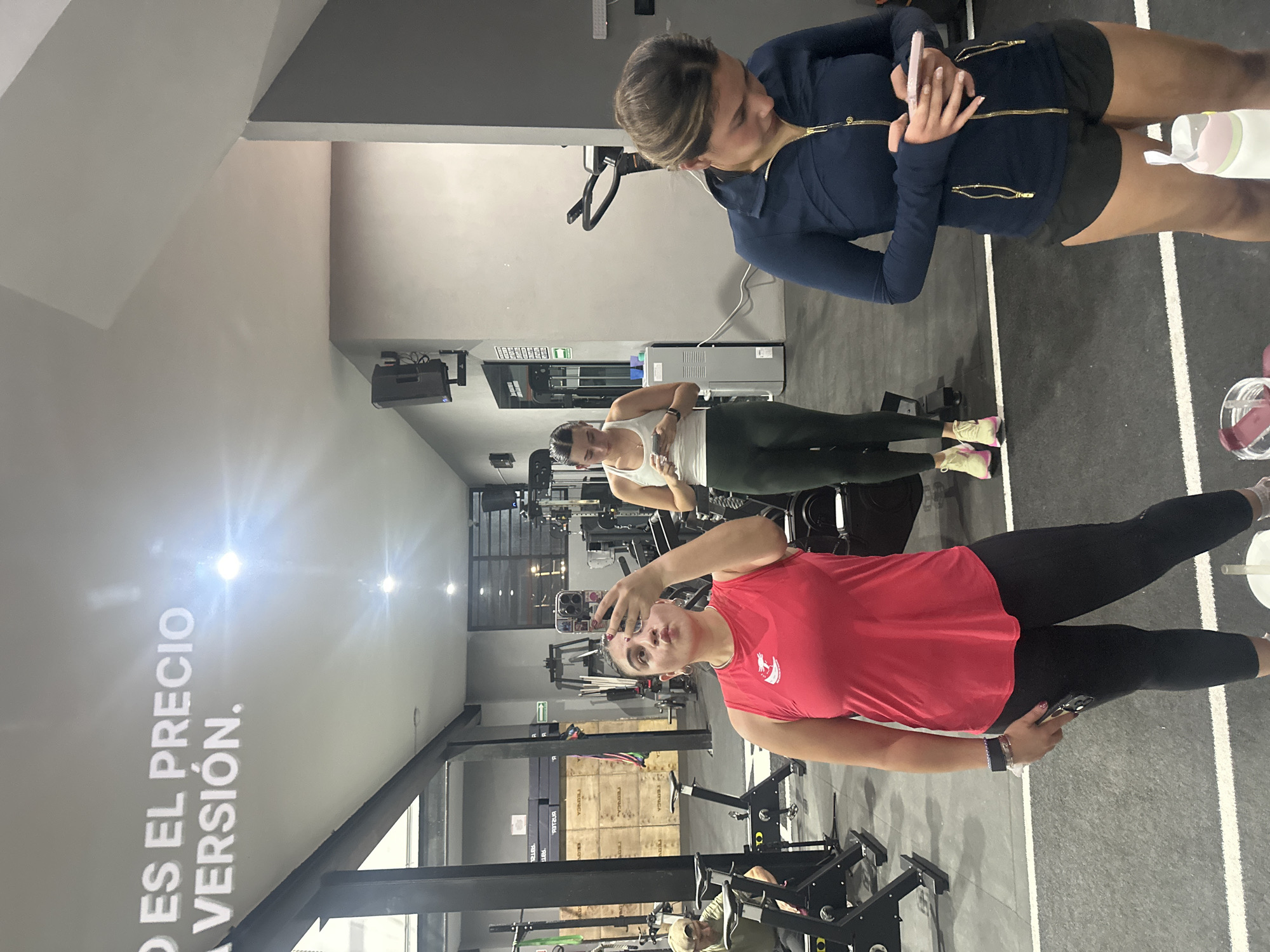 The image captures a scene inside a gym located in San Bernardino Tlaxcalancingo, Mexico. Three women are present. One woman is taking a selfie, dressed in a red tank top and black leggings. A second woman is standing to the left, wearing a white tank top and dark green leggings. The third woman stands off to the right, wearing a navy blue zip-up jacket and black shorts, looking at her phone. Gym equipment is visible in the background. The gym has a grey color scheme with a motivational phrase printed on the wall.