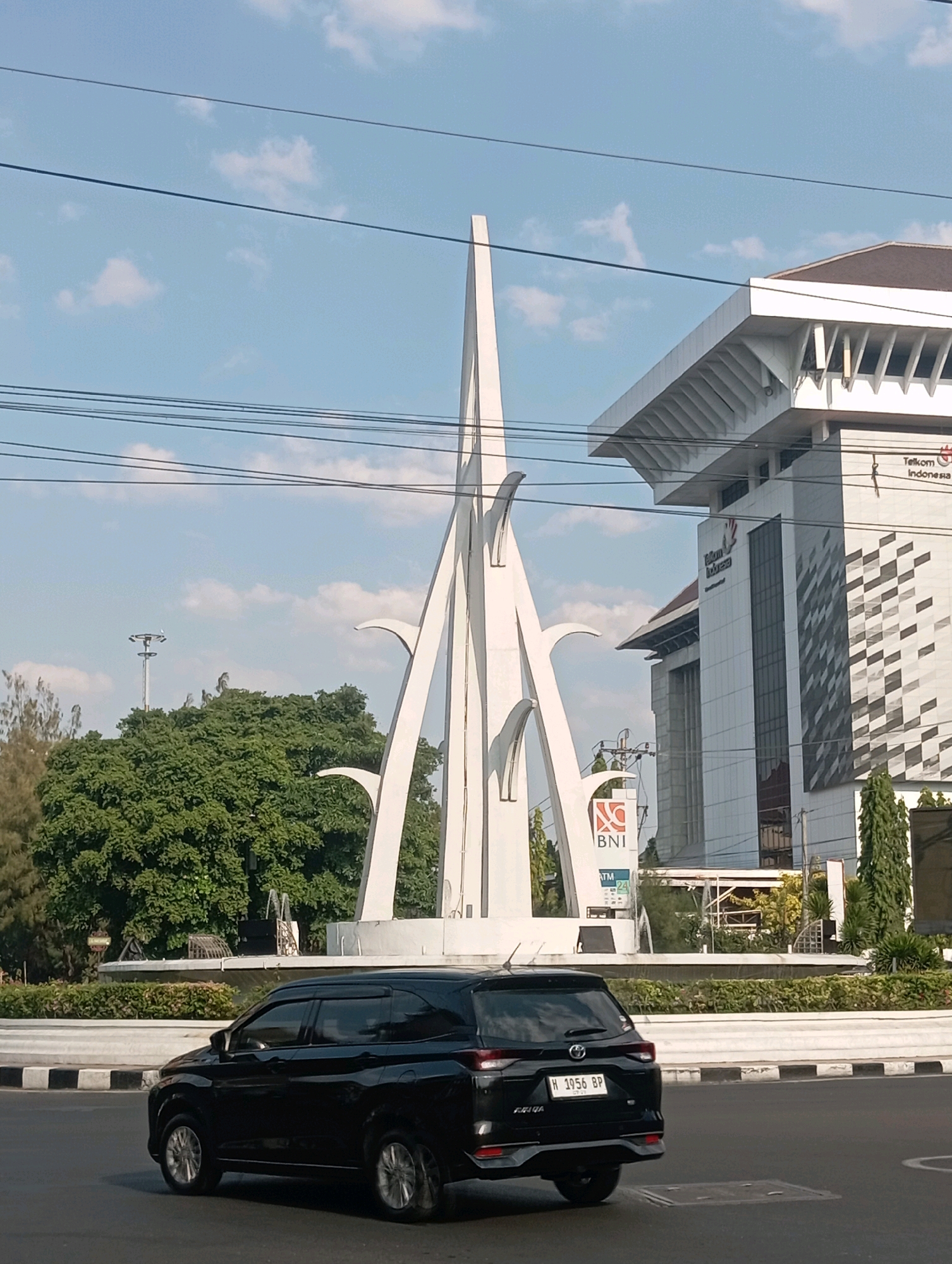 In Semarang, Indonesia, a black SUV is captured in motion on a paved street, its rear facing the camera. The vehicle displays a license plate reading "H 1956 BP". Behind the car, a modern roundabout features a tall, white abstract sculpture as its centerpiece. To the right of the sculpture stands a multi-story building with a white facade, adorned with the "Telkom Indonesia" logo. In front of this building, a smaller sign displays "BNI" and "ATM". Lush green trees form a dense backdrop to the roundabout, and overhead, a network of electrical wires crisscrosses the clear blue sky dotted with white clouds. The scene is bathed in daylight.