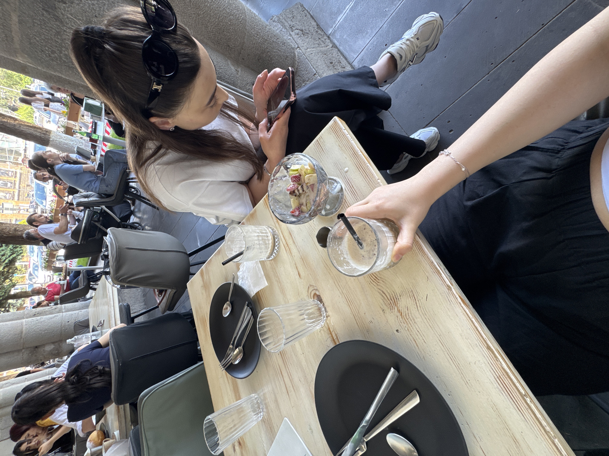 The image depicts an outdoor seating area of a cafe in Gyumri, Armenia. A wooden table is set with plates, cutlery, and glasses. Two women are visible at the table, one of whom is looking at her phone. A fruit salad is in a glass bowl on the table. In the background, other patrons are seated at tables, and trees and buildings are visible. The overall scene suggests a casual daytime gathering at a cafe.