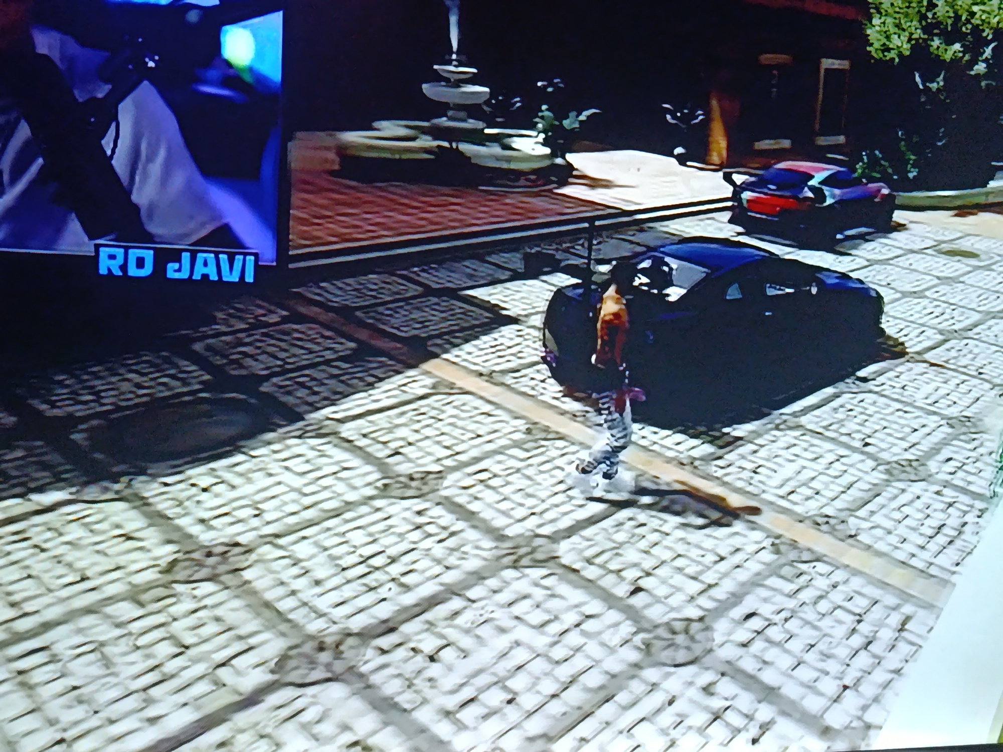 The image shows a video game scene on a monitor. In the foreground, there is a person standing next to a dark-colored car. Behind the car, there is another car with red and purple accents. In the background, there is a tiered fountain, greenery, and a building facade. The game interface displaying "RO JAVI" is visible in the lower left corner of the monitor. The ground is paved with interlocking square stones. The location is not discernible as Juan Griego, Venezuela without additional information.