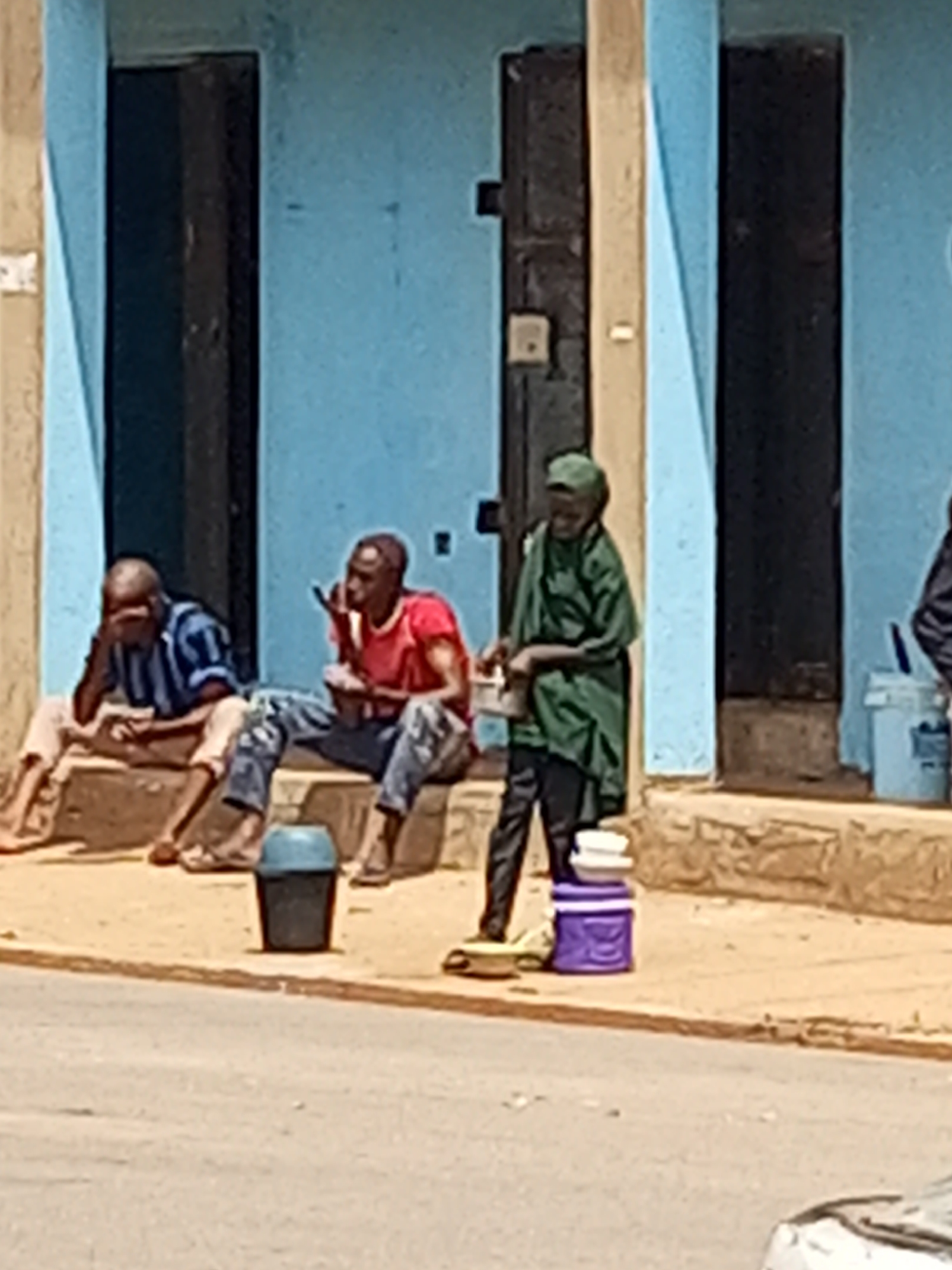 In Tempure, Nigeria, three individuals are present outside a building with a light blue facade and dark doorways. On the left, a person wearing a striped blue and white shirt and light-colored pants sits on a raised step, facing left with their hand on their forehead. To their right, another person in a red t-shirt and blue jeans sits on the same step, holding a mobile phone and appearing to be in conversation. A third person, on the right, wears a green headscarf and a green outfit, standing and holding a white container. In front of the building, a dark blue bin with a light blue lid is placed on the ground. Further to the right, a purple container with white lids is stacked on the ground. The ground in front of the building appears to be a mixture of sand and pavement, with a paved road in the foreground.