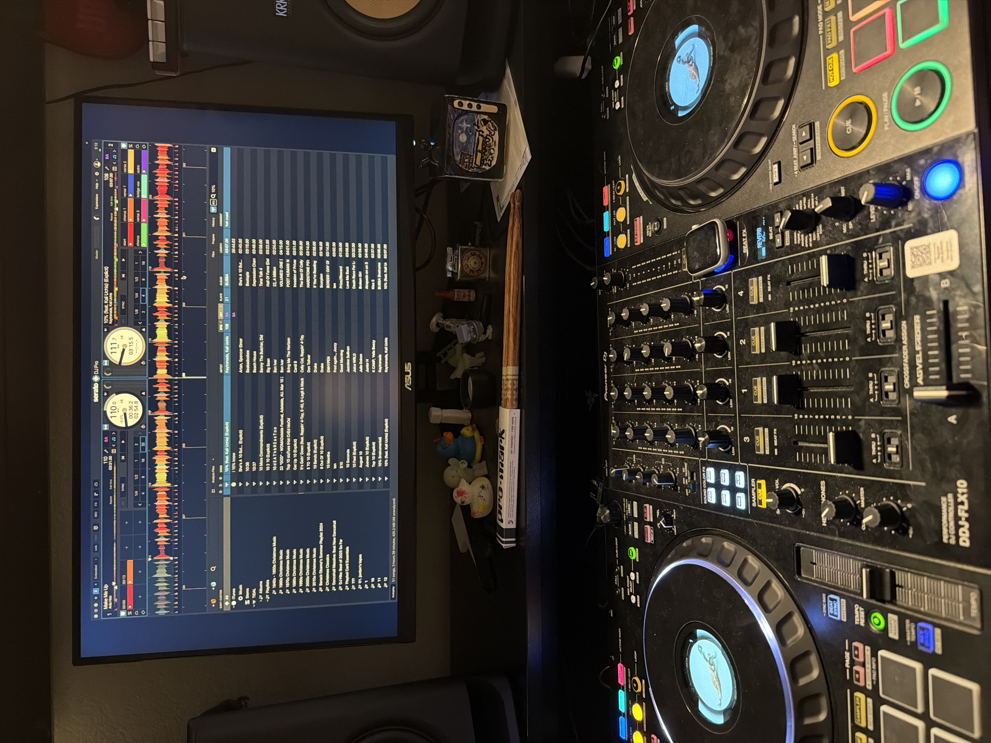 The image captures an indoor DJ setup from a high-angle perspective, showing a computer monitor, a DJ controller, and a studio monitor. The scene is illuminated by artificial light, suggesting an evening or nighttime setting.
The main subjects are the DJ equipment. On the left, a large monitor displays the Serato DJ Pro software interface, showing waveforms, track information, playlists, and a song titled "10% (feat. Kali Uchis) (Explicit)" by Kaytranada. Key elements like BPM (111.7 and 110.0), elapsed time (00:36.2, 02:54.8, 03:15.5), and track details are visible on the screen, which also bears the "ASUS" brand logo.
To the right, a Pioneer DDJ-FLX10 performance DJ controller dominates the frame, with numerous illuminated buttons, knobs, and faders. An Apple Watch rests on one of the jog wheels. The controller features sections labeled "PAD MODE," "HOT CUE," "CUE," "PLAY/PAUSE," "BEAT FX," "REVERB," "SAMPLER," "MASTER," and "TEMPO." A "KRK" brand studio monitor is partially visible in the upper background.
On the desk between the monitor and the controller, various small items are arranged, including a drumstick labeled "pro-mark," several small figurines or toys, and a decorative box. The overall impression is a dedicated home studio or DJ workspace.