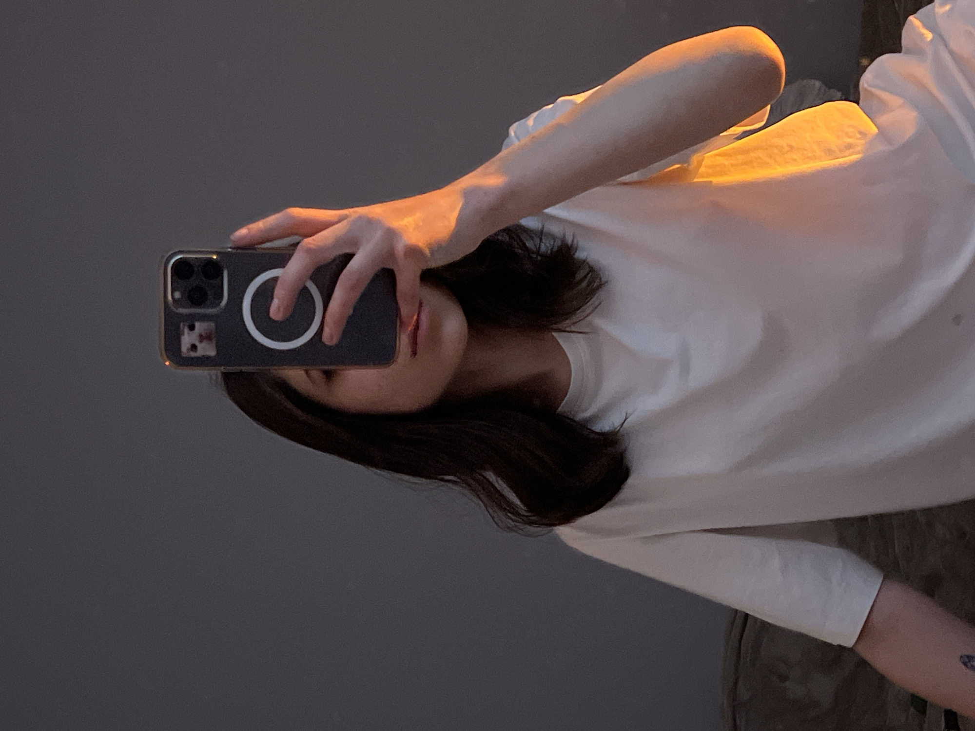 A person with shoulder-length dark hair is depicted in a medium shot, holding a dark-colored smartphone to partially obscure their face, revealing only the chin and lips. The individual wears a plain white short-sleeved t-shirt. A small, dark tattoo is visible on the inner left forearm. The smartphone, likely an iPhone due to the triple camera array, features a white circular MagSafe ring on its dark case, which also displays a small image of a cat's face in the upper left corner. The person holds the phone with their right hand, which, along with the right arm and portions of the t-shirt, is strongly illuminated by a warm, orange-yellow light source originating from the right side of the frame. This lighting creates prominent highlights and shadows. The background is a plain, dark grey wall or surface, suggesting an indoor environment. A textured, dark fabric object, possibly upholstery, is partially visible in the lower left foreground. The image provides no identifiable visual information to determine its geographical location in Ternopil, Ukraine, or any other specific city.