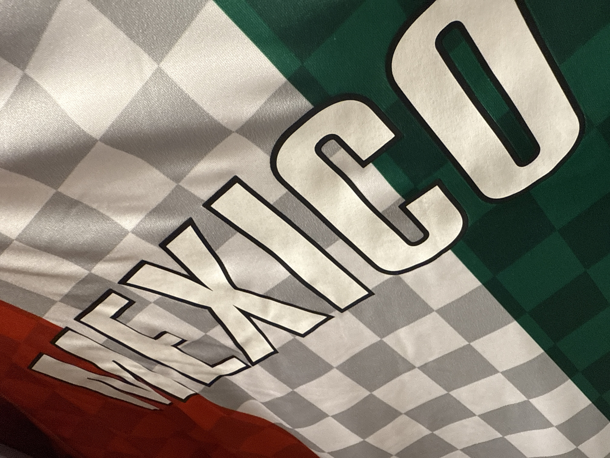 A close-up shot of a "Mexico" jersey. The fabric is divided into three sections, red, white and green, with white and gray checkered patterns on the white section. The word "Mexico" is printed diagonally across the fabric in large, bold letters with black outlines.