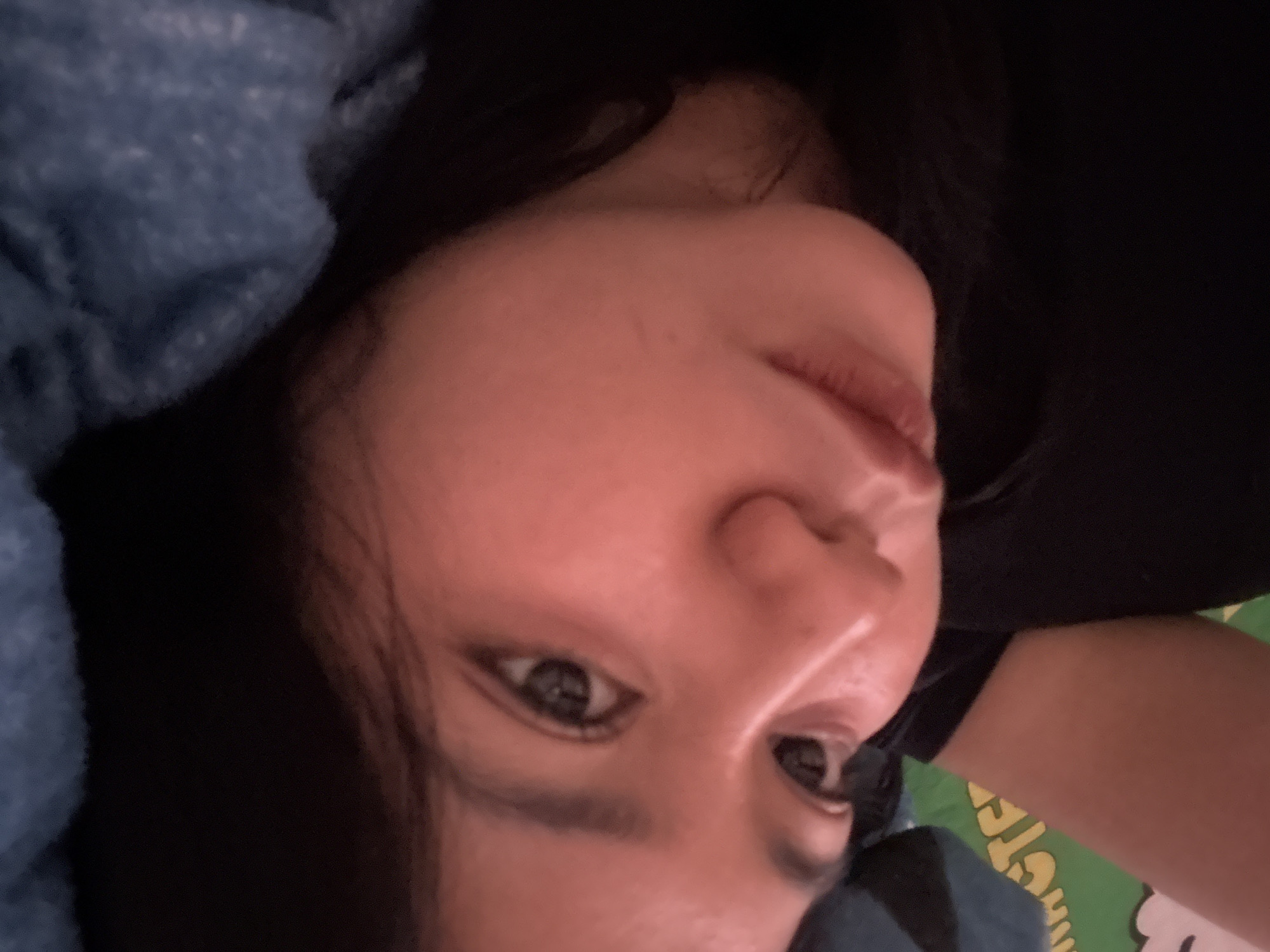 The image is a close-up, upward-angled portrait of an adult female, lying down and looking directly at the camera. Her face dominates the frame, tilted diagonally. She has dark hair, and her expression is calm with a subtly relaxed mouth, while her eyes, defined by visible eyeliner, gaze towards the viewer.
She is dressed in a dark top, with a textured, possibly knitted, blue fabric (likely a blanket or garment) partially covering her on the left side. In the background on the lower right, a green fabric, possibly a blanket or sheet, is visible, bearing partial yellow text. Although indistinct, parts of the text appear to show "GOTE" followed by other letters and a partial "R" below, possibly forming a word like "GOTCHA!" or a brand name.
The setting appears to be indoors, likely in a bedroom or living area in North Jakarta, Indonesia, given the casual pose and soft furnishings. The dim lighting casts a warm, reddish tone on her skin, suggesting an evening or low-light environment. The overall ambiance is relaxed and intimate.