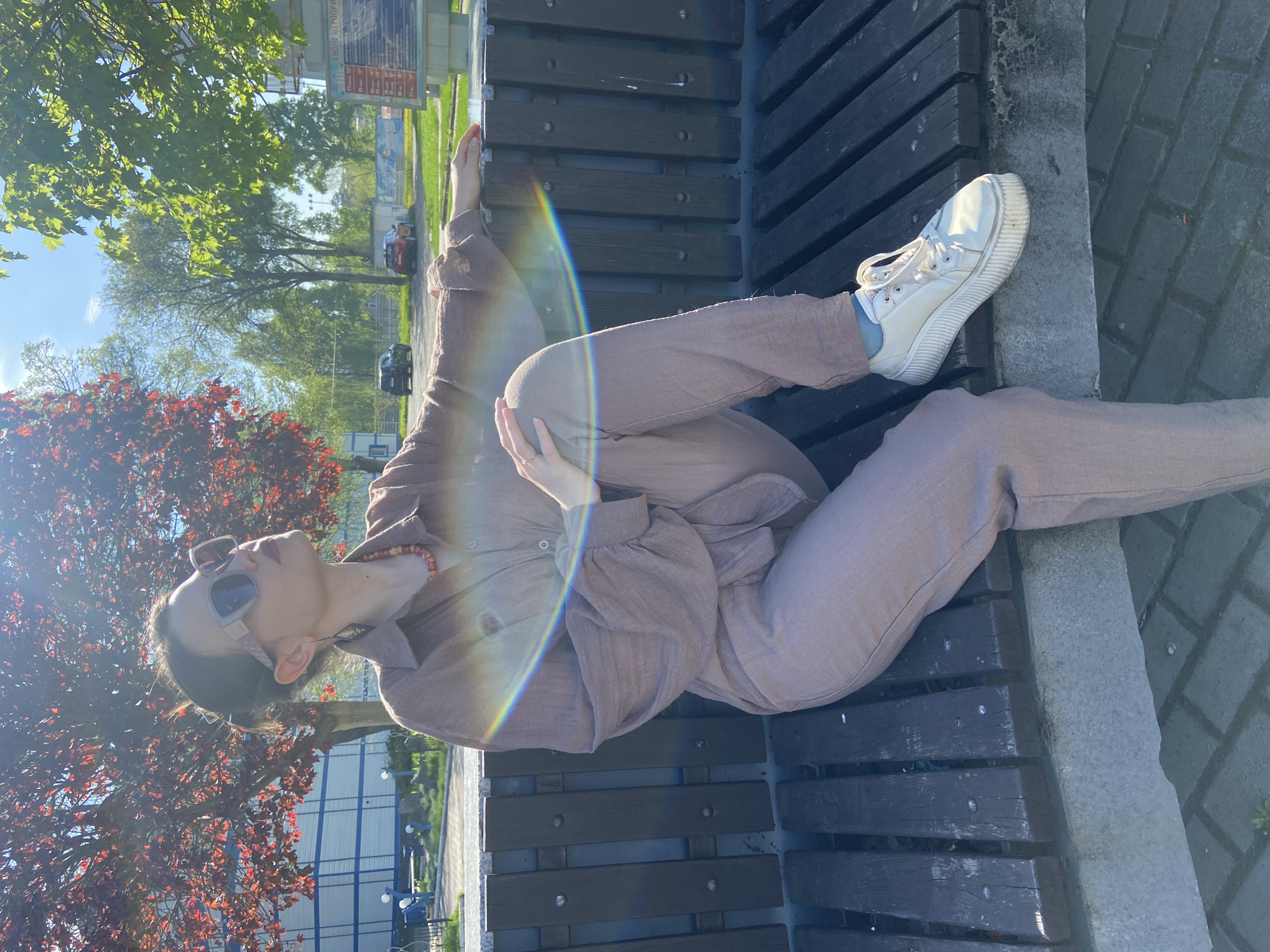 A person in sunglasses and a brown outfit is reclining on a park bench. The person is wearing white sneakers and a necklace. A tree with red leaves is to the left of the person. The bench has dark-colored wooden slats with a gray stone border. In the background are green trees, buildings, and cars. The location is the city of Chaikovsky, Russia.