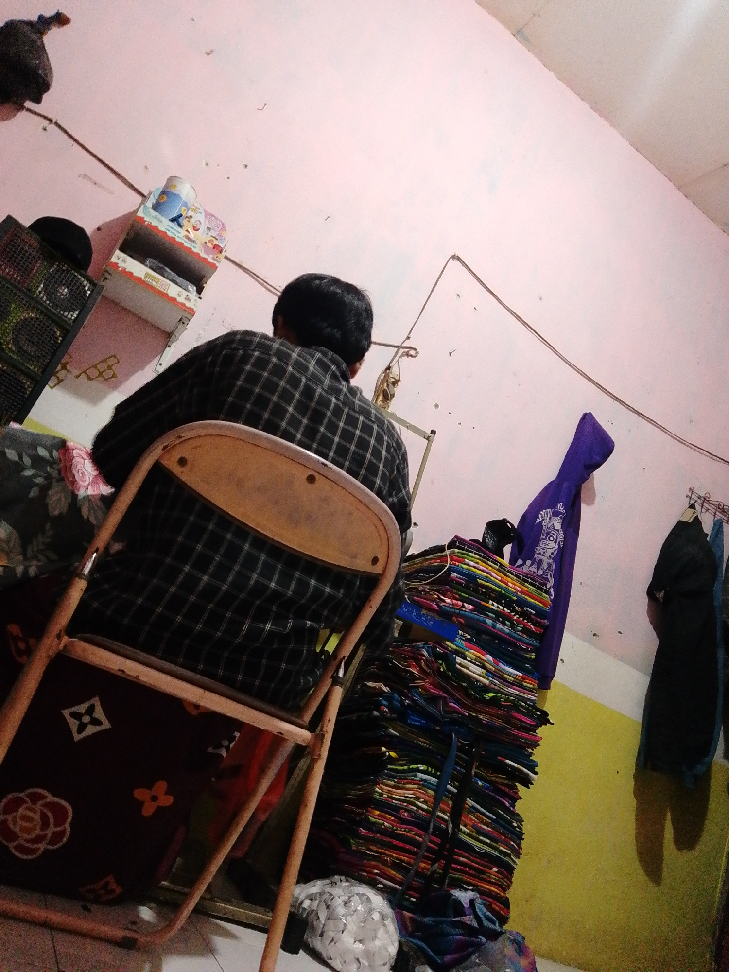 In the city of Bohar, Indonesia, a person is seated on a folding chair facing away from the camera, wearing a black and white checkered shirt. The chair has a wooden seat and backrest, and is positioned in front of a stack of colorful fabric items. Behind the person, a light pink wall is visible, adorned with electrical wires and various objects. To the left, a dark-colored speaker or electronic device sits on a shelf, with a black cap on top. Further to the left, a shelf contains a few items, including what appears to be a toy or figurine. To the right of the fabric stack, a purple hooded garment hangs on the wall. The floor appears to be tiled or concrete. The overall scene suggests an indoor setting, possibly a residence or a small shop.