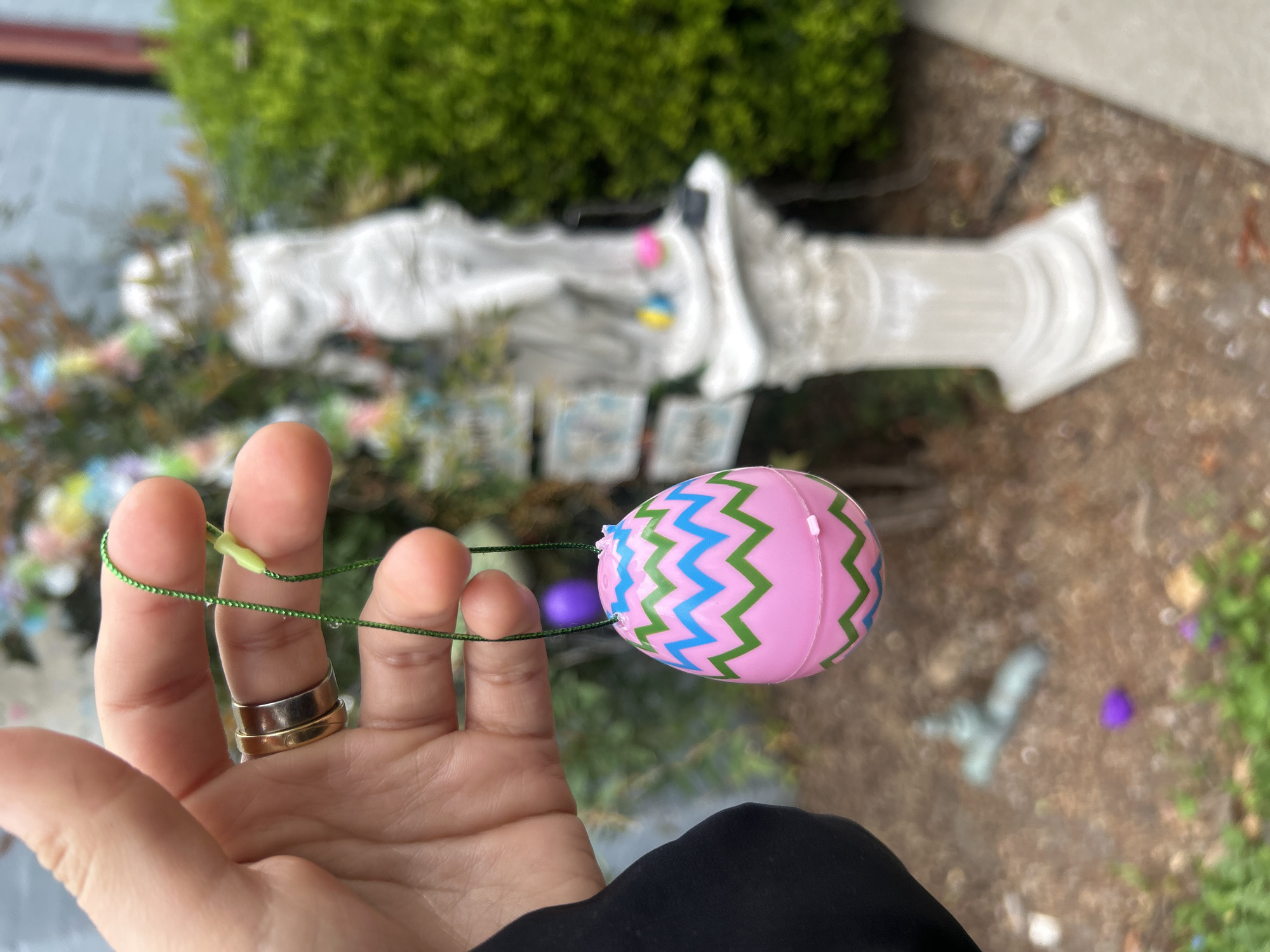 A hand holding a pink plastic Easter egg with green and blue zigzags is in focus. The egg is suspended by a green string wrapped around the fingers of the hand, which also has two gold rings. The background is out of focus but appears to be a yard or garden area with greenery, statues, and small signs. The statue in the background appears to be a broken religious statue on a column. There is foliage in the background including bushes and various plants. The location is difficult to determine with certainty, but the style of the yard and the light suggest a residential area. The overall feel is one of casual observation of an Easter decoration.