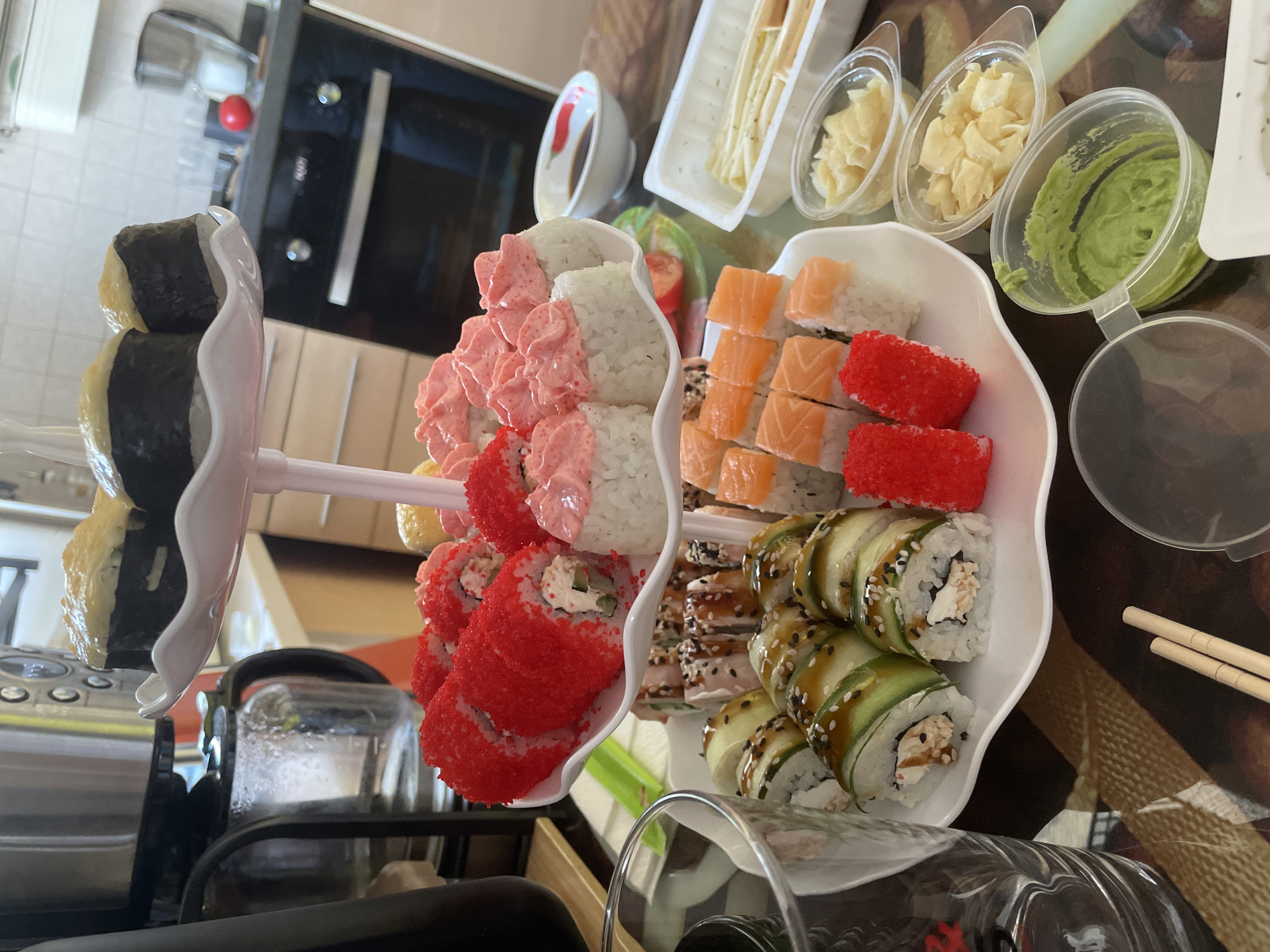 The image shows a two-tiered white serving dish filled with sushi, along with accompanying condiments, on a dark, patterned tabletop. The top tier contains maki rolls with pink topping, tobiko-covered rolls, and tamagoyaki. The bottom tier has rolls with salmon and tobiko, and eel-cucumber rolls topped with sesame seeds. Condiments including wasabi, pickled ginger, and soy sauce are visible in plastic containers nearby. Chopsticks are present. The setting appears to be indoors, likely a private residence in Perm, Russia, given the user's stated location. Other visible objects include a wine glass, a kettle, and part of a kitchen appliance. No people are present.