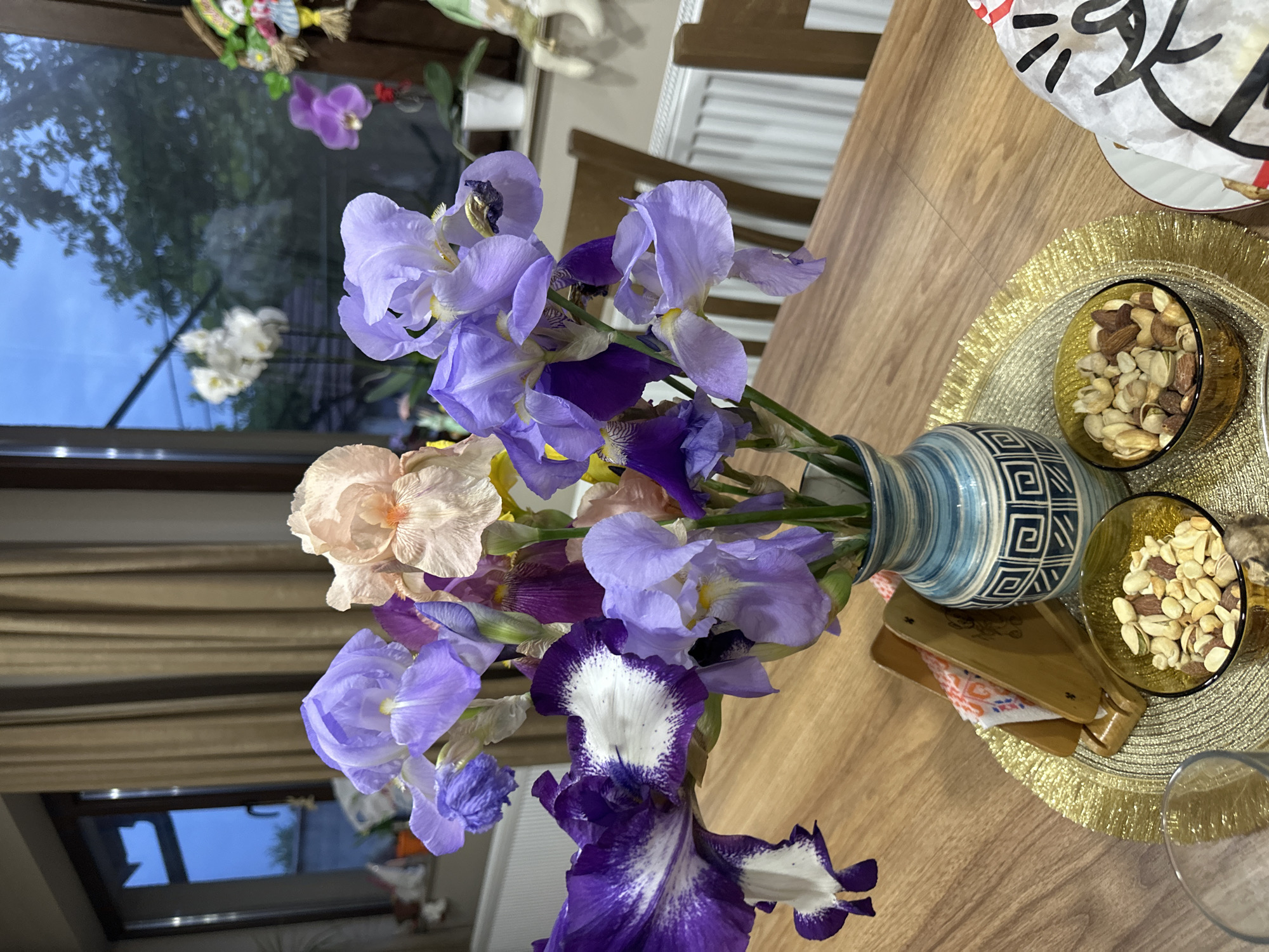 The image depicts a bouquet of irises in a blue and white patterned vase placed on a wooden table. A small bowl of mixed nuts sits beside the vase on a gold placemat. A paper napkin and a pair of wooden chopsticks are also present on the table. Through a window in the background, glimpses of trees and the sky can be seen, along with other floral decorations. The location is Lehchevo, Bulgaria.