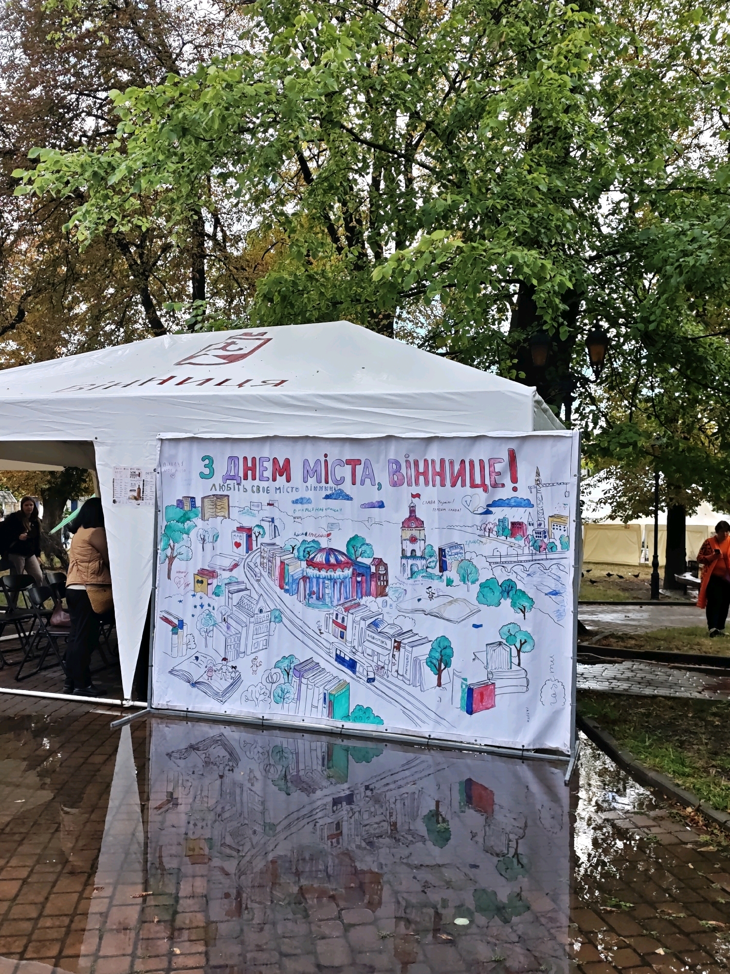 A large white tent is set up outdoors, likely in a park or public square in Vinnytsia, Ukraine. The tent's fabric has a red logo with some writing, partially obscured by trees. A prominent banner, attached to the front of the tent, features a colorful hand-drawn mural depicting a city scene with buildings, trees, a carousel, and people reading. The mural is titled "З ДНЕМ МІСТА, ВІННИЦЕ!" which translates to "Happy City Day, Vinnytsia!". Below this, in smaller text, it says "ЛЮБИТЬ СВОЄ МІСТО ВІННИЦЮ!" meaning "Loves its city of Vinnytsia!". There are also smaller handwritten notes and drawings on the banner.
The ground in front of the banner is wet and reflective, indicating recent rain, with brick paving visible beneath the water. Several people are visible. To the left, two women stand near chairs, one wearing a brown jacket and the other a dark jacket and pants. To the far right, a person in an orange garment walks away from the camera. Several pigeons are gathered on the wet pavement in the background. The surrounding environment includes lush green trees with some autumn foliage, suggesting the photograph was taken in the fall. The overall impression is of an outdoor event celebrating the city.