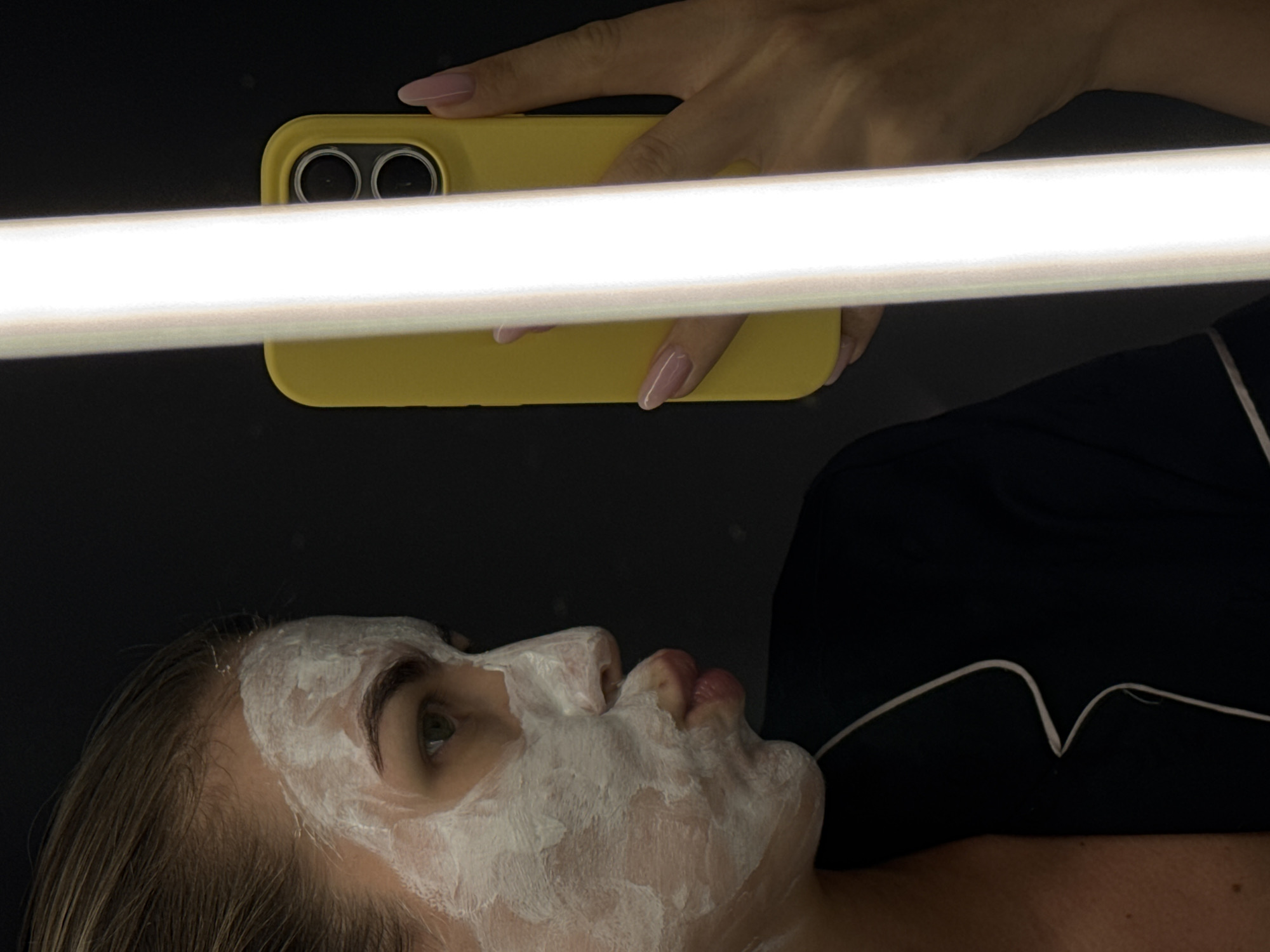 A person with a white facial mask is lying down. Above the person, a hand holds a yellow cell phone. A white light source is positioned between the cell phone and the person's face. The background is dark. The location is unknown.