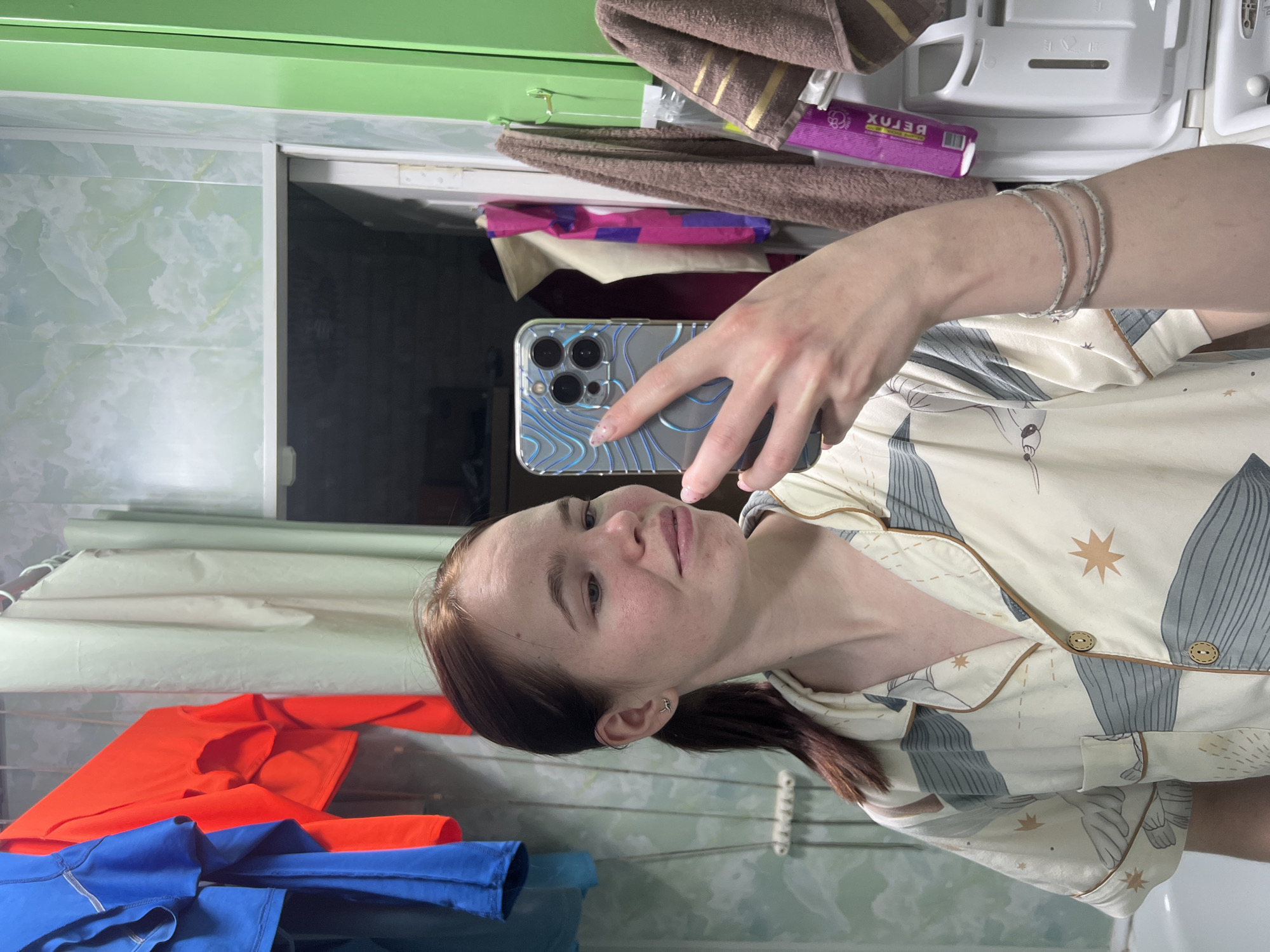 A young woman is captured in a selfie taken in a bathroom or dressing area, likely in Chaikovsky, Russia. She is shown from the chest up, with her head tilted slightly. Her light brown hair is pulled back into a ponytail. She wears a cream-colored pajama top adorned with a pattern of greyish-blue abstract shapes, possibly whales or fish, and brown starbursts, featuring brown piping and wooden buttons. On her left wrist, she wears several thin, silvery bangles. She holds a smartphone with a distinctive clear case featuring a blue and silver wavy pattern, its three camera lenses visible. Her expression is calm, with a slight smile.
The background reflects a domestic setting. To her right, a green shelf or cabinet is visible, with brown towels draped over it. Below the towels, various toiletries and products are stored, including a purple box with the mirrored text "XULƎЯ" (RELAX). White shelving units are also present. To her left, bright orange and blue long-sleeved clothing items are hanging on a drying rack. The walls or tiles in the background have a light green, marble-like pattern. The overall scene suggests a casual, indoor moment, possibly at home.