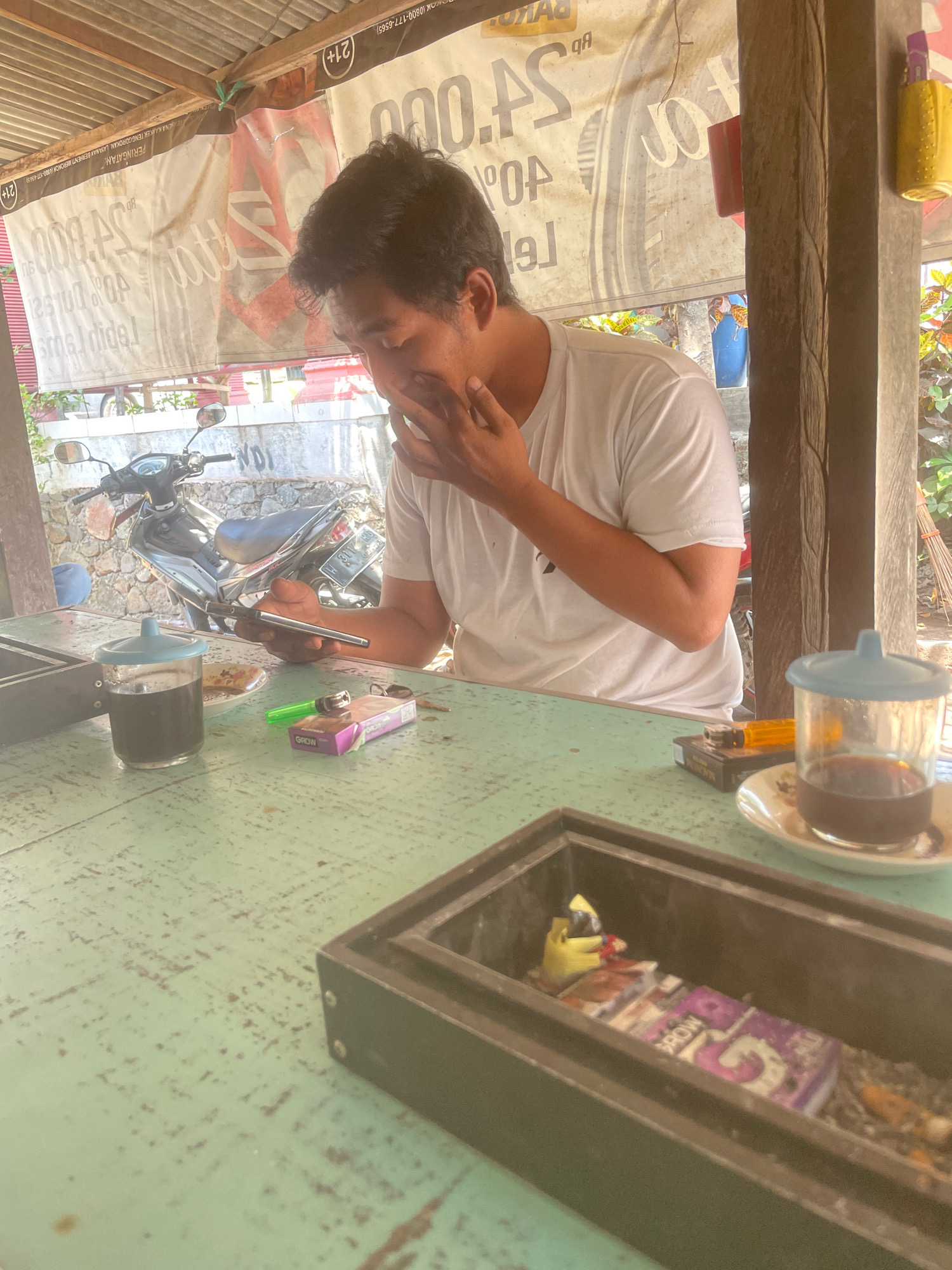 A man is sitting at a table outdoors under a corrugated metal roof. He is wearing a white t-shirt and has his hand to his face, appearing to be thinking or rubbing his eyes. On the green-painted wooden table in front of him are a glass of dark liquid, a cigarette pack, a lighter, and a pen or stylus. A motorcycle is parked in the background, and banners with text are hanging behind the man. The overall setting suggests a casual, possibly roadside establishment in Kebon Waris, Indonesia, given the presence of Indonesian text and the commonality of such establishments in the region.