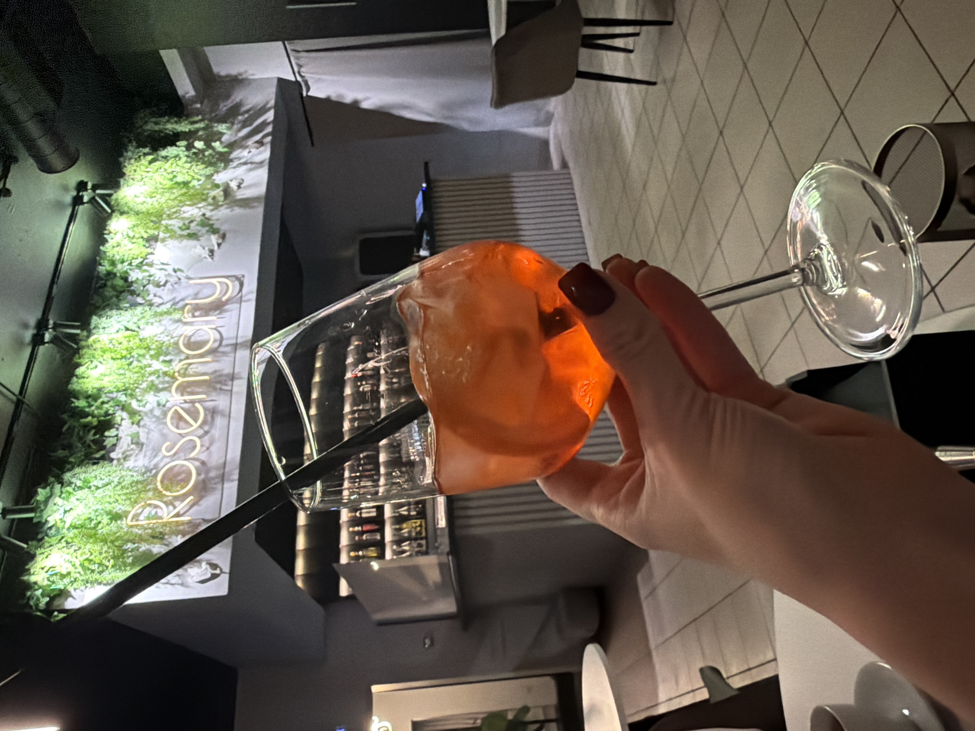 An interior shot depicts a modern bar or restaurant named "Rosemary" in Votkinsk, Russia. The foreground shows a person's left hand, with dark red manicured fingernails, holding a stemmed glass filled with an orange beverage, ice cubes, and a citrus slice, along with a black straw. The glass is positioned slightly above a small, dark, cylindrical bin on the floor.
In the midground, a bar counter with a ribbed facade is visible, behind which shelves display numerous bottles of alcohol. Above the bar, a white structure adorned with illuminated artificial green foliage features a glowing yellowish-white neon sign spelling "Rosemary." The ceiling is dark with exposed pipes and track lighting.
To the far left, a portion of a white table and a white cup and saucer are present. To the right, a table and a single dark-backed chair are visible, with white curtains partially obscuring a wall. The floor is tiled with light-colored square tiles and dark grout. The scene is generally dimly lit, with strategic illumination on the sign and foliage.