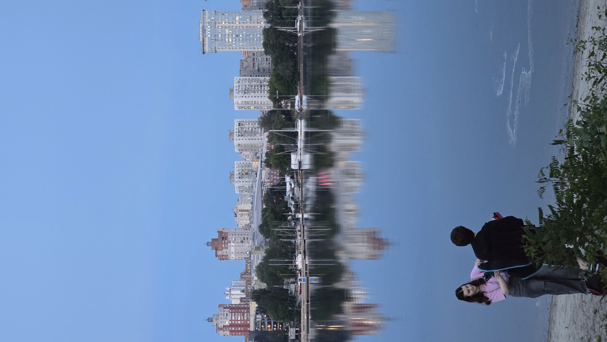 This image depicts a twilight scene along the Dnieper River in Kyiv, Ukraine, with a view towards a section of the city's residential skyline, likely the Obolon district.
The upper third of the frame shows a clear, light blue sky. The central and lower portions are dominated by the calm, reflective surface of the Dnieper River, appearing dark blue-grey. It prominently mirrors the city skyline above. Lighter, frothy patterns are visible on the water's surface near the right foreground shore.
A dense residential skyline extends horizontally across the horizon. On the far left, a multi-story building with a reddish-brown facade and an ornate, multi-tiered dark roof structure is visible. To its right, a series of multi-story apartment blocks, predominantly white or light grey, are present, interspersed with green trees. On the far right, a tall, slender, modern glass high-rise building with visible internal lights stands prominently. Below the buildings, along the riverbank, a marina or yacht club is evident, featuring multiple sailboats and motorboats docked along a pier. Some faint red light reflections are visible in the water beneath the marina area.
In the bottom left foreground, a sandy bank slopes gently towards the water. Two individuals are present on this bank. The person on the left is a female with dark hair, wearing a light pink short-sleeved top and dark grey trousers. She faces right, looking towards the other individual, and holds a dark blue object, possibly a carrying case or bag. Her expression is a slight smile. The person on the right is male, positioned with his back largely to the viewer, facing the water. He wears a dark, possibly black, hooded jacket and dark trousers. He holds a red object in his left hand. A cluster of green leafy bushes partially obscures the view of the water in front of and to the right of the individuals.