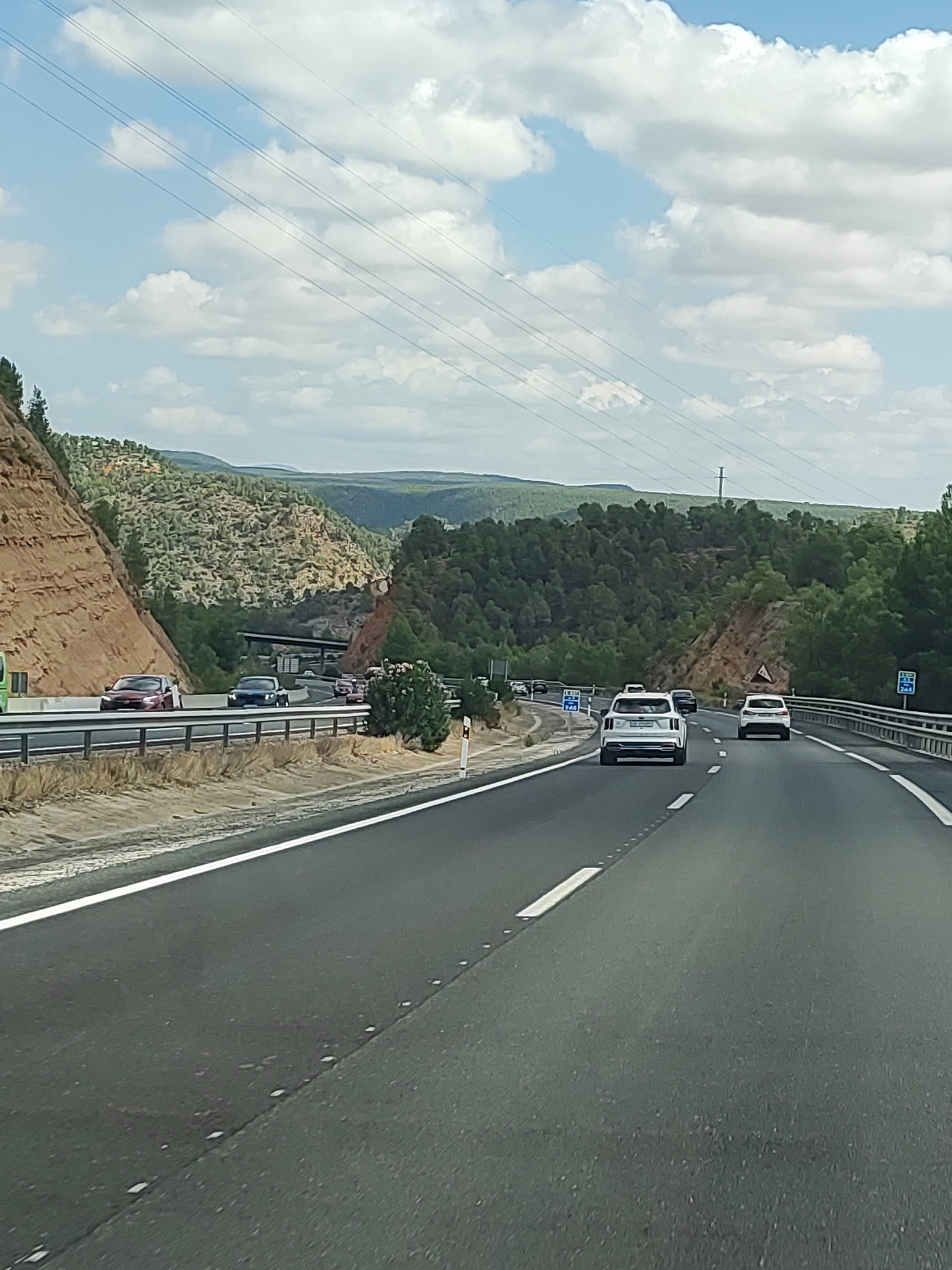 The image shows a highway scene, likely taken from a moving vehicle, in the vicinity of Minglanilla, Spain. The road is asphalt and divided into multiple lanes by white lines. Several cars are visible on the highway, including a white SUV in the foreground, a black car behind it, and other vehicles further ahead. On the left side of the highway, there is a steep, rocky embankment with some vegetation. On the right side, the highway is bordered by a metal guardrail and beyond that, a landscape of rolling hills covered in dense green trees and some scattered bushes. Power lines are visible crisscrossing the sky, which is partly cloudy with a bright blue sky peeking through. The overall environment suggests a rural or semi-rural mountainous area.