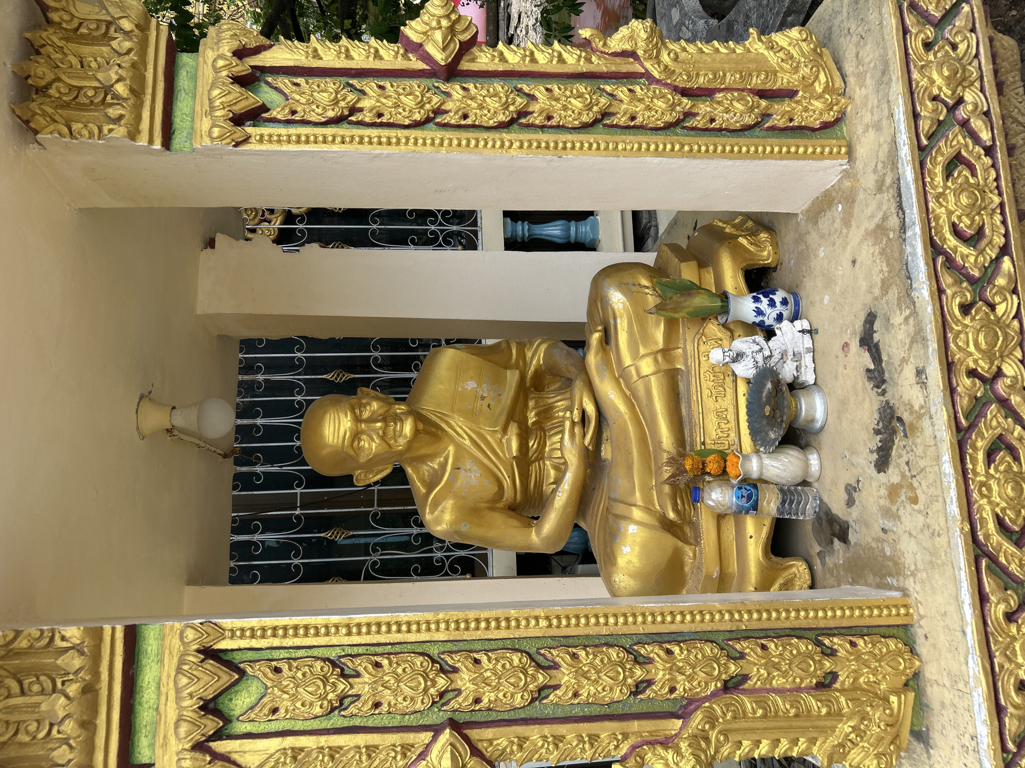 A golden Buddha statue is seated within a shrine adorned with ornate gold carvings. The statue is positioned centrally, with a white figurine and a blue and white vase to its right. A bottle of water and some offerings are placed in front of the statue. The shrine itself is decorated with detailed gold motifs in green and red accents. The background shows a barred window and a blue pillar. The shrine is situated outdoors, with a hint of greenery and what appears to be a body of water in the background, suggesting a location in Ko Samui District, Thailand.