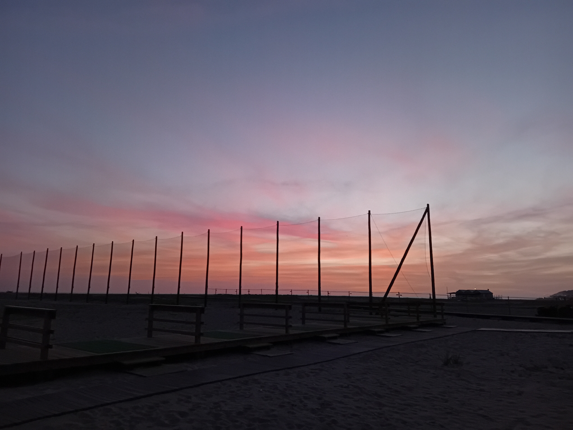 The image depicts a serene twilight scene, likely at either sunrise or sunset, over a sandy expanse that appears to be a golf driving range. The sky is the most prominent feature, displaying a striking gradient of colors. Just above the distant horizon, vibrant hues of orange and pink glow brightly, gradually transitioning upwards into soft purples and finally into a deep, muted blue at the very top of the frame. Delicate, wispy clouds are illuminated by these colors, adding texture to the sky.
In the midground, a long line of tall, dark poles stands vertically, supporting a fine-mesh net stretched between them. This structure forms a protective barrier typical of a golf driving range, designed to contain golf balls. These poles and the net are silhouetted against the colorful sky, indicating the dim light of twilight.
The foreground consists of dark, sandy ground, suggesting a beach or a large, open sandy area. Several wooden platforms, which serve as tee boxes for golfers, are visible on this sandy surface. Some of these platforms include small wooden benches on either side, indicating individual practice bays.
In the far distance on the right side of the frame, a small, dark building with a discernible roofline is silhouetted against the lighter part of the sky, possibly a clubhouse or a beachside structure. The horizon line where the land meets the sky is subtly visible.
No individuals are present in the image. The absence of people, combined with the tranquil colors of the sky, creates a calm and contemplative atmosphere. The scene captures a moment of natural beauty at dusk or dawn. There is no visible text in the image.