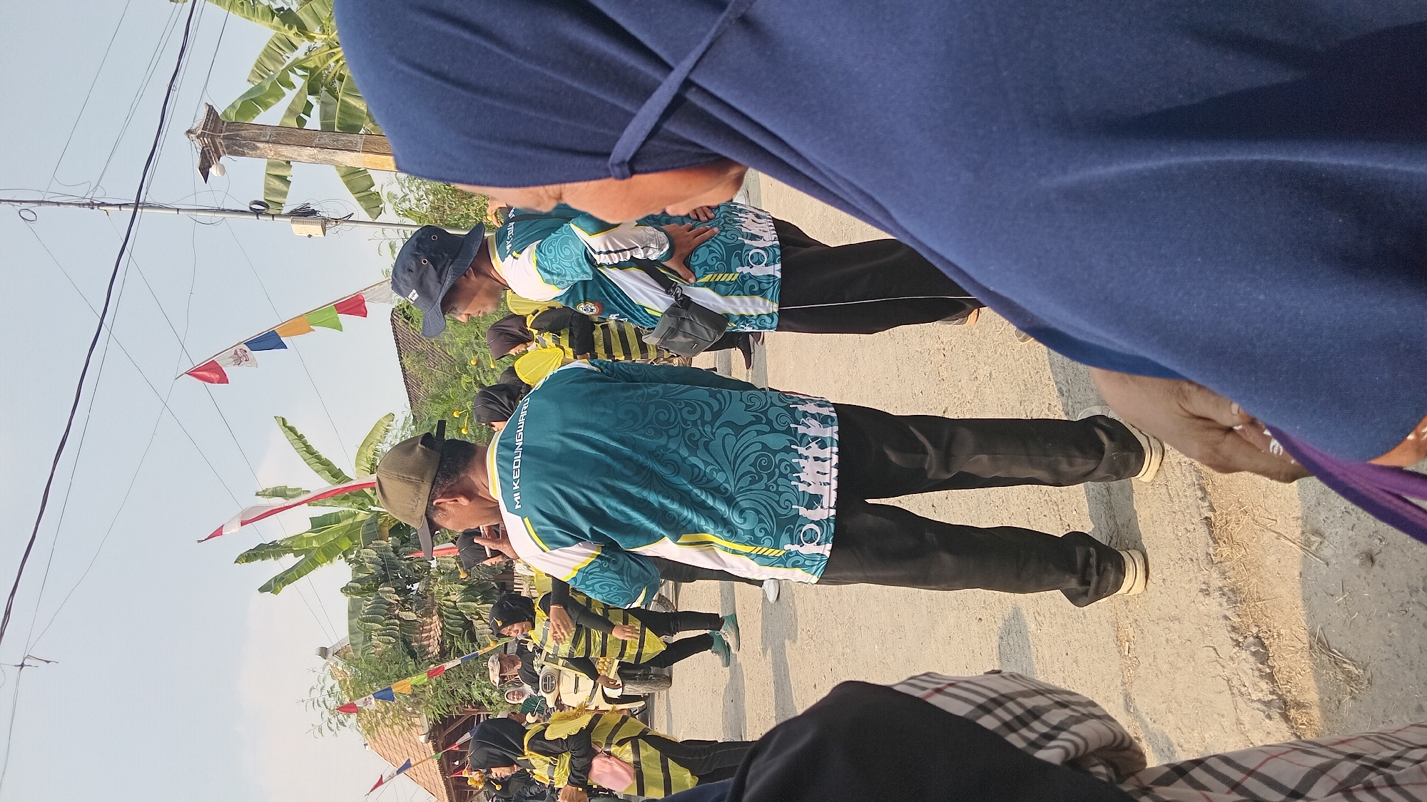 The image captures a street scene in Kedungwaru, Indonesia, likely during a procession or outdoor event. In the foreground, a person wearing a blue hijab is partially visible, looking towards the right. Behind them, two men in matching teal and white shirts with a patterned design and black pants stand facing away from the camera. The man on the left wears a green cap, while the man on the right wears a dark blue bucket hat. Both shirts have text that reads "MI KEDUNGWARU."
Further in the background, several individuals dressed in yellow and black bee-themed costumes are visible. Some are holding yellow objects, possibly wings or props. A white scooter is parked behind this group. The environment suggests a warm, sunny day, with clear skies and some vegetation, including banana trees. Decorative flags, including Indonesian national flags (red and white), are displayed on poles. The ground is unpaved and appears to be a dirt or gravel road. The overall atmosphere is festive and communal, suggesting a local celebration or parade.