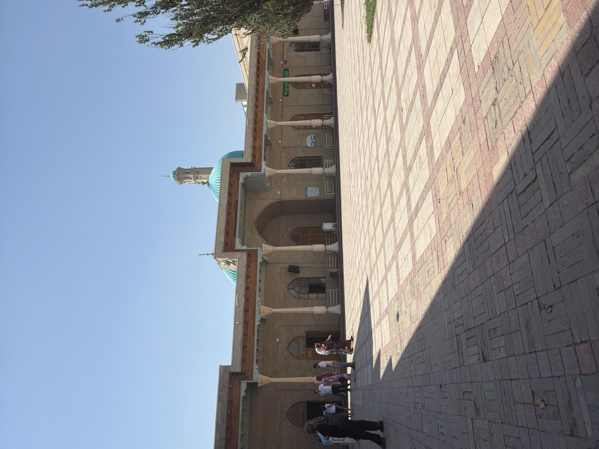 This image depicts the exterior of the Zangi-Ota Mosque complex in Zangiata, Uzbekistan, under a clear blue sky. The foreground features a vast, paved courtyard with a patterned arrangement of light and darker rectangular tiles, intersected by a prominent shadow cast by an unseen object to the left. Several individuals, some appearing to be tourists and others possibly locals, are distributed across the courtyard, with a group of people near the entrance of the mosque and a single person in dark attire standing to the far left.
The mosque itself is constructed of light brown brickwork, characterized by a long, colonnaded porch with a wooden overhang supported by numerous white pillars. The porch features arched entrances and windows with wooden frames. Above the porch, a light-colored roofline is visible. A prominent turquoise-colored dome and a minaret, also with turquoise accents, rise above the mosque's structure, indicating its religious significance. To the right of the mosque, a tall, leafy green tree stands against the sky. A digital display showing "PESHIN 13:00" is mounted on the mosque's facade above the entrance. Informational signs are also visible near some of the entrances. The overall scene suggests a public space, likely a place of worship and historical interest, with visitors engaged in exploration or prayer.