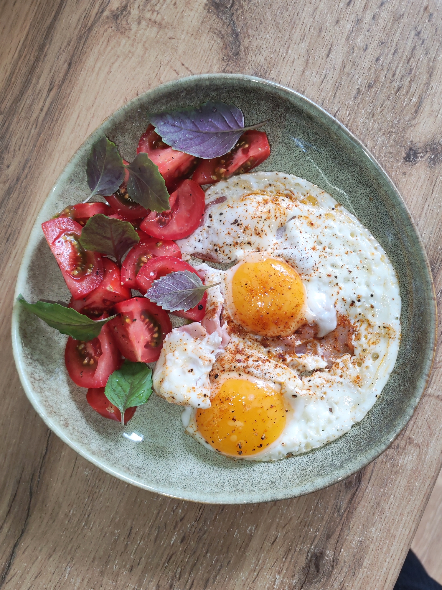 A top-down, close-up shot captures a ceramic plate holding a prepared meal situated on a light brown wooden surface. The round plate, featuring a textured greenish-grey glaze, contains two fried eggs with visible, bright orange yolks and cooked whites, which are sprinkled with dark pepper flakes and reddish-orange spices. Adjacent to the eggs, on the left side of the plate, is a portion of sliced red tomatoes, cut into wedges, appearing fresh and moist. Several small, whole basil leaves, a mix of green and purple varieties, are scattered among the tomatoes and lightly over the eggs. The wooden surface beneath the plate exhibits a distinct, slightly diagonal grain pattern. No individuals or additional environmental context are visible to indicate a specific geographic location, such as the city of Izhevsk, Russia.
