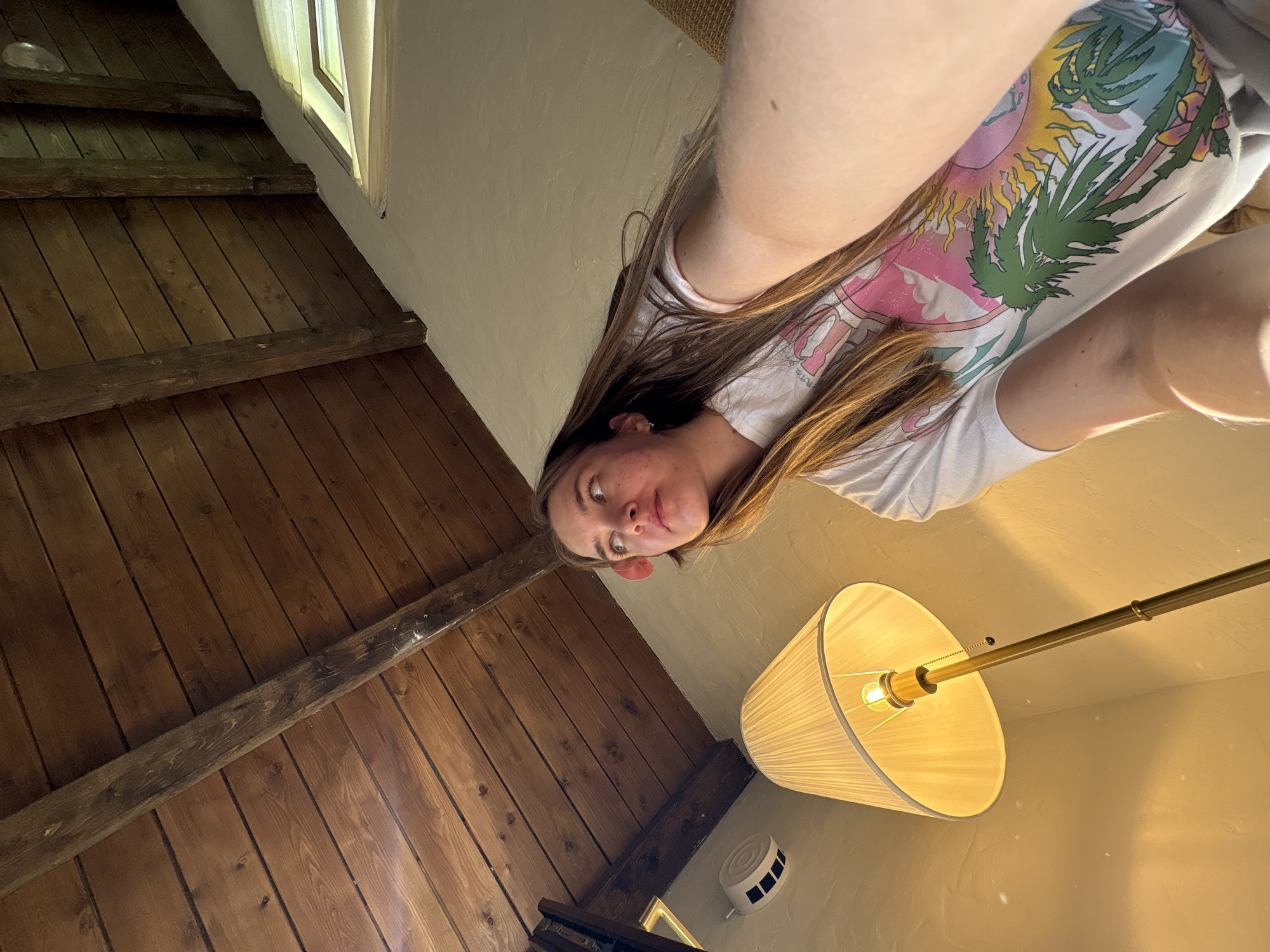 A tilted perspective photograph depicts a person with long brown hair, wearing a white t-shirt with a graphic print, positioned against a backdrop of a wooden ceiling and a wall with a lamp. The wooden ceiling is composed of planks, some with visible knots. The lamp has a white pleated shade and a brass-colored stand. A white circular object with black rectangular vents is visible on the wall. The photograph does not indicate that it was taken in Sibir', Russia.