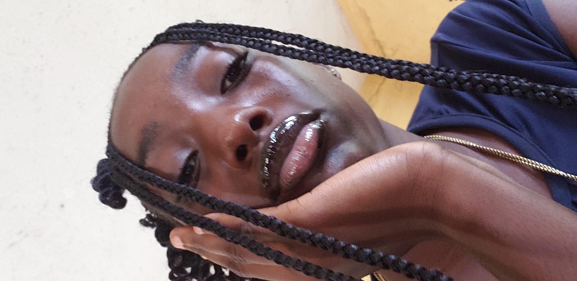 This close-up, diagonally oriented portrait features a person with rich, dark skin, likely female, posing against a plain, light-colored wall. The subject's head is tilted to the right, and she rests her chin on her left hand, which subtly supports her jawline. Her eyes are open, looking towards the camera with a calm or contemplative expression. Her lips are slightly parted and adorned with a dark, glossy finish that catches the light. Her facial features are well-defined, and her skin exhibits a healthy, subtle sheen, particularly on the forehead and nose.
Her hair is styled in numerous neat, dark box braids that frame her face and extend both upwards and downwards out of the frame. She is wearing a dark blue garment, possibly a shirt, which is visible on her upper right side. A delicate gold chain is also visible around her neck, resting against her clothing. The background is a simple, light beige or off-white wall with a subtle textured appearance, suggesting an indoor or shaded outdoor setting. The lighting is soft and natural, indicating it is daytime. The image appears to be a direct, posed shot. While no specific location cues are visible within the image, the context provided is Kaduna, Nigeria. No visible text is present.