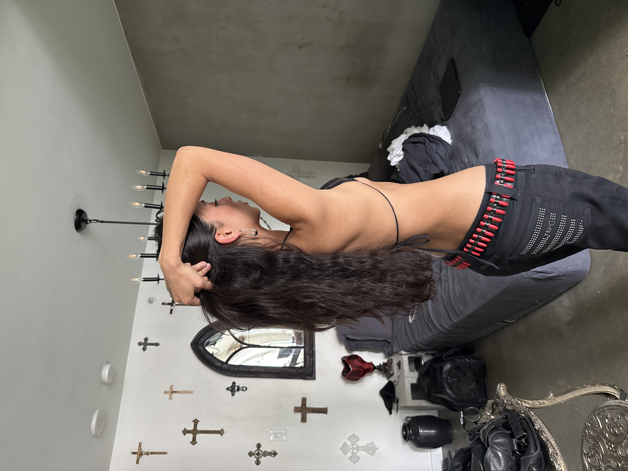 An indoor, side-profile shot captures a person with long, dark, wavy hair, posing in a room with a distinctive aesthetic. The person wears a black string top and black pants, accessorized with a prominent red and black studded belt resembling a bullet belt. Their left arm is raised, with hands near their head, seemingly running fingers through their hair. Their face is seen in profile, with eyes closed or looking downwards, conveying a calm or contemplative expression. A silver earring is visible in their left ear.
The room features light grey walls and a darker grey floor, possibly concrete. A black chandelier with exposed bulbs hangs from the ceiling. One wall is decorated with multiple cross ornaments of various sizes and materials, along with an arched, dark-framed mirror. To the right, a dark grey bed or large couch is partially visible, with some clothes and a tablet on it. In the bottom right, an ornate silver table or chair with intricate carvings adds to the decor. Other objects include a black backpack, a black jug or urn, and a red, sculptural decoration on a surface. A white smoke detector is mounted on the wall. No visible text is present. The overall setting suggests a personal space with a gothic or alternative style, potentially in Vancouver, Canada.