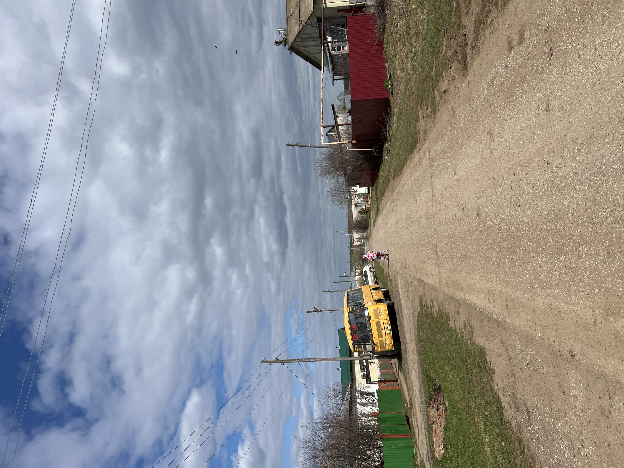 The scene is a dirt road in Перевозное, Russia, flanked by vegetation and houses. A yellow school bus is parked on the left side of the road, and a small figure wearing pink clothing is walking away from the bus further down the road. Overhead, utility poles line the road, with wires stretched across the sky. The sky is filled with a mix of blue sky and cloud cover.